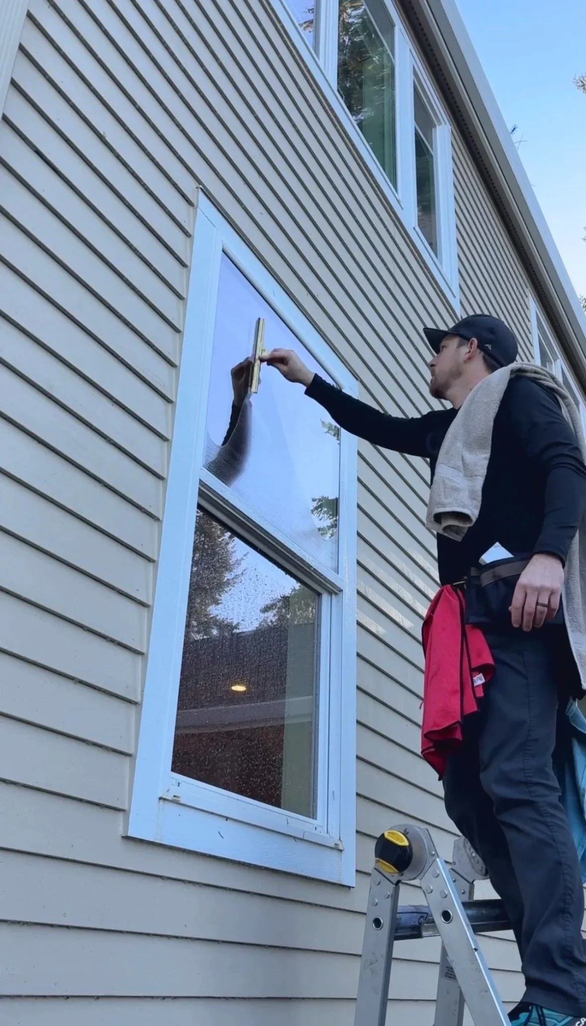 A man standing on a ladder cleaning a window on the exterior of a house with beige siding using a squeegee.
