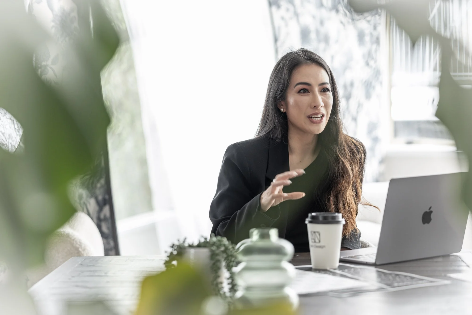 A woman with long dark hair speaking while sitting at a desk with a laptop, coffee cup, and decorative items, in a bright room with large windows and blurred greenery in the foreground.