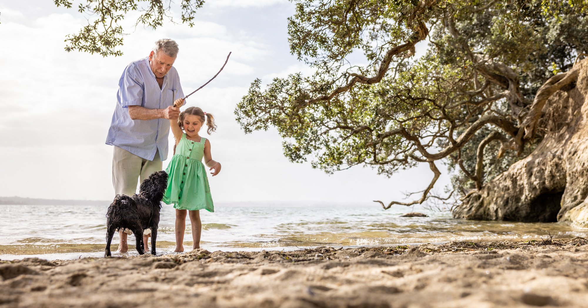 An elderly man, young girl, and a black dog on a beach, with the man holding a stick and the girl smiling as she stands near the dog in shallow water, with trees and rocks along the shoreline.