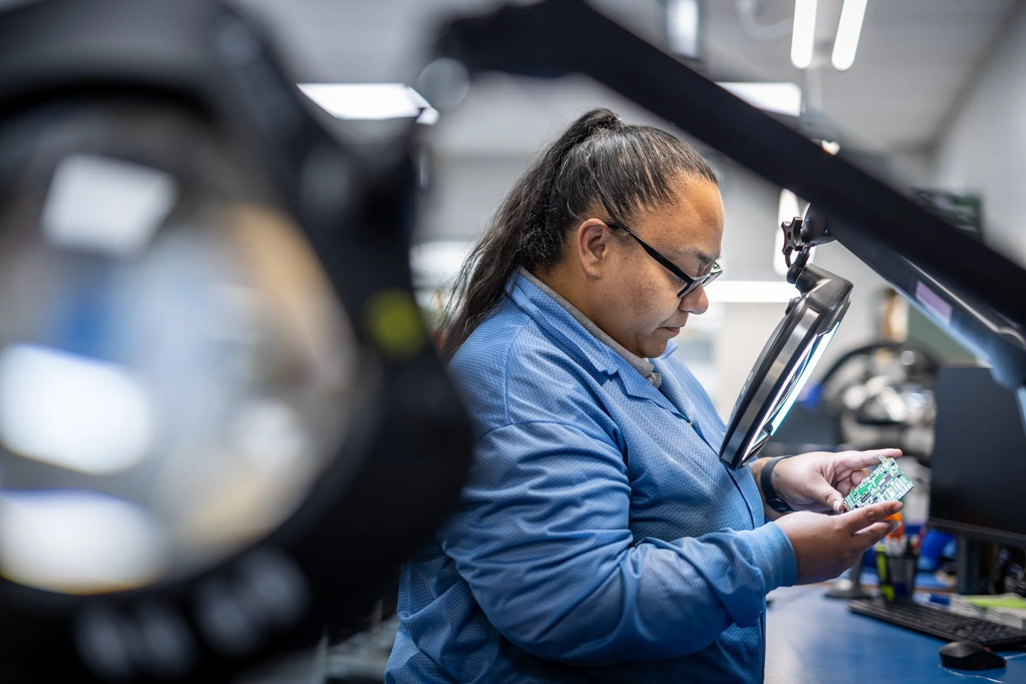 A person wearing glasses and a blue lab coat working on a circuit board at a workstation, with electronic equipment and components visible in the background.  Photographed by Alex Wallace Photography