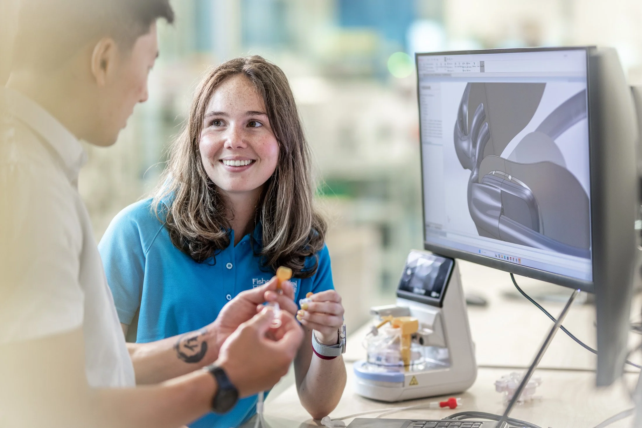 Two people working in a laboratory, one woman smiling and looking at the other person who is holding a small object. They are sitting at a desk with a computer displaying 3D modeling or engineering design.