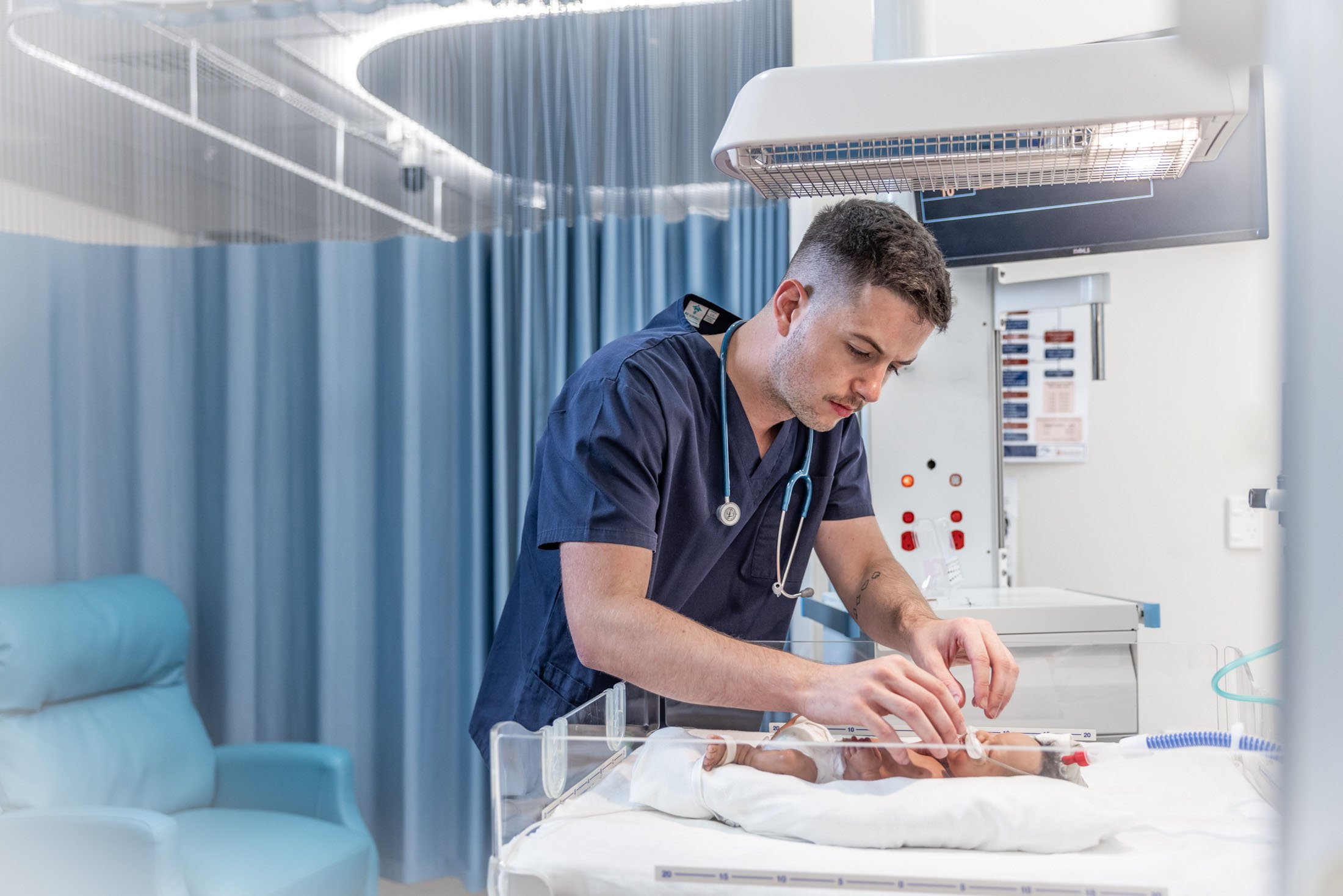 A male healthcare professional, wearing navy scrubs and a stethoscope, tending to a newborn baby in a hospital incubator.