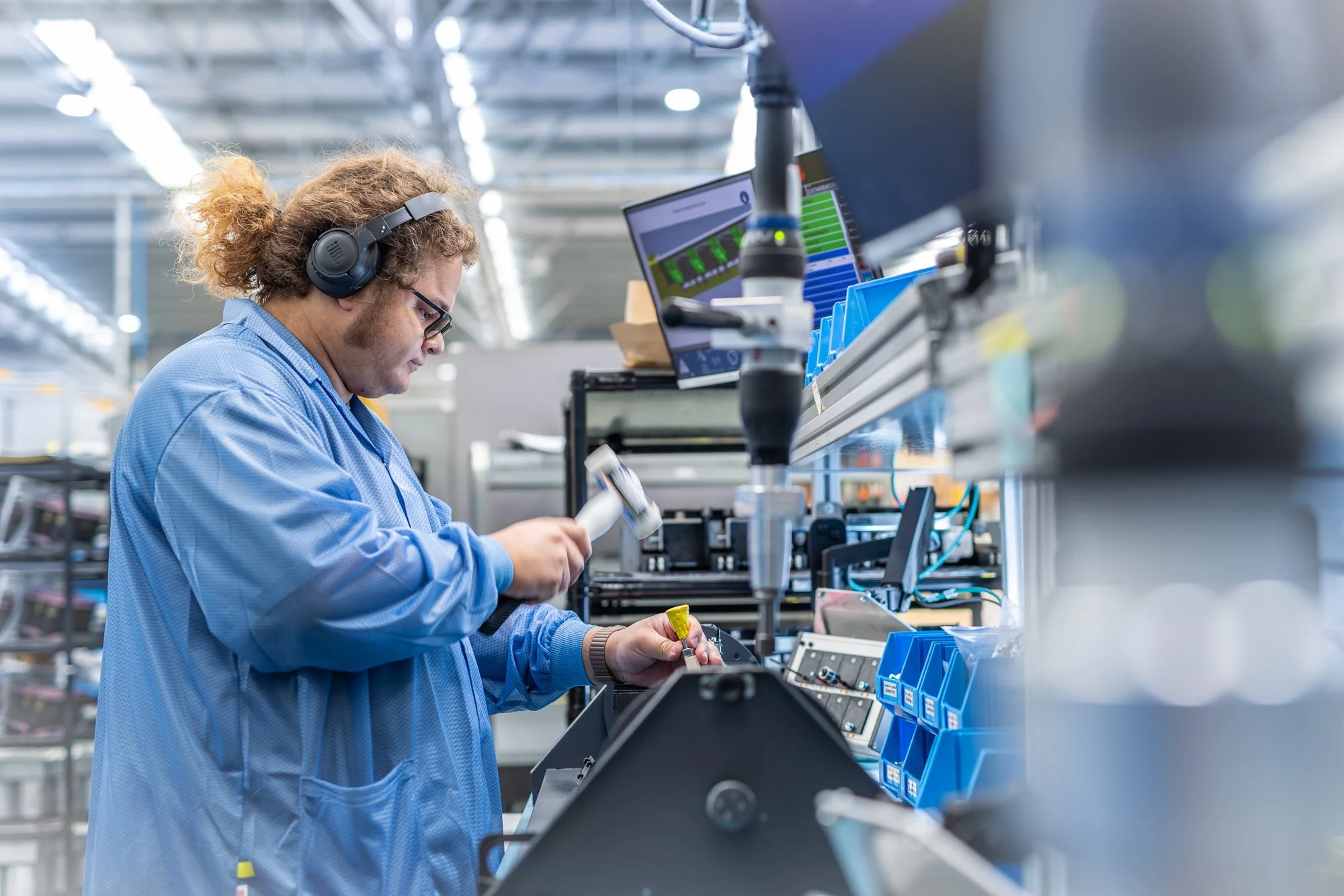 A male factory worker with curly hair in a ponytail, wearing glasses, black headphones, a blue lab coat, and handling a small tool in a manufacturing or electronics assembly environment.  Photographed by Alex Wallace Photography