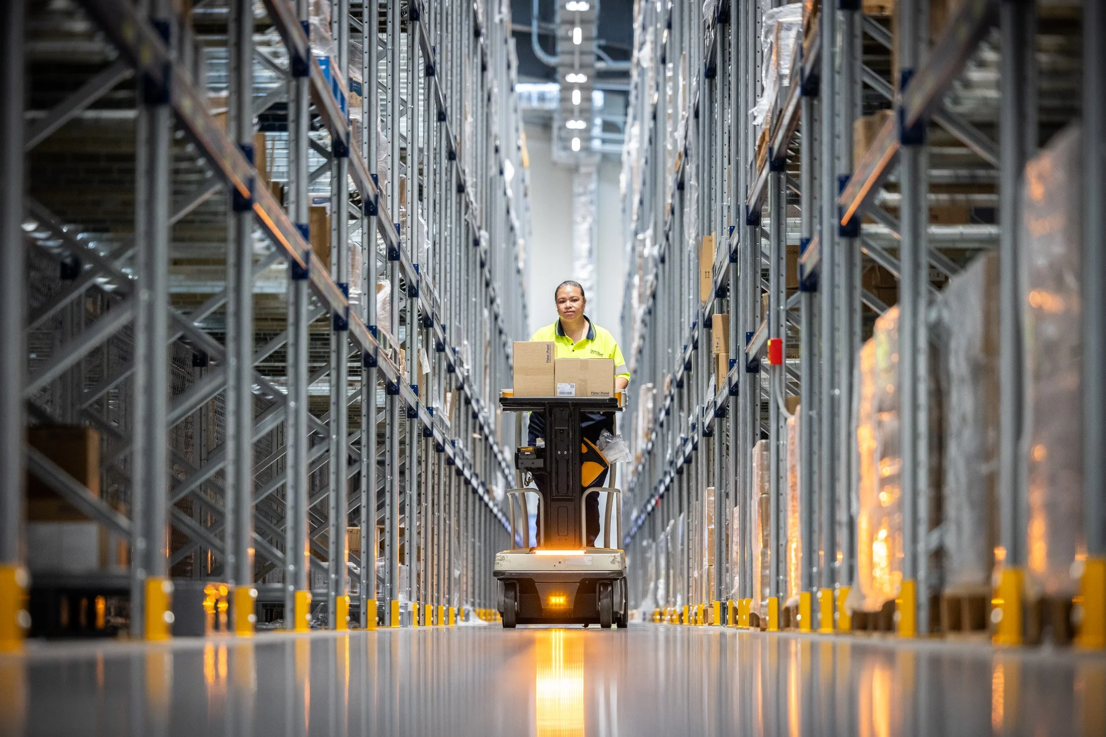 A warehouse worker in a high-vis shirt operating a robotic cart among tall metal shelves filled with boxes. Photographed by Alex Wallace Photography