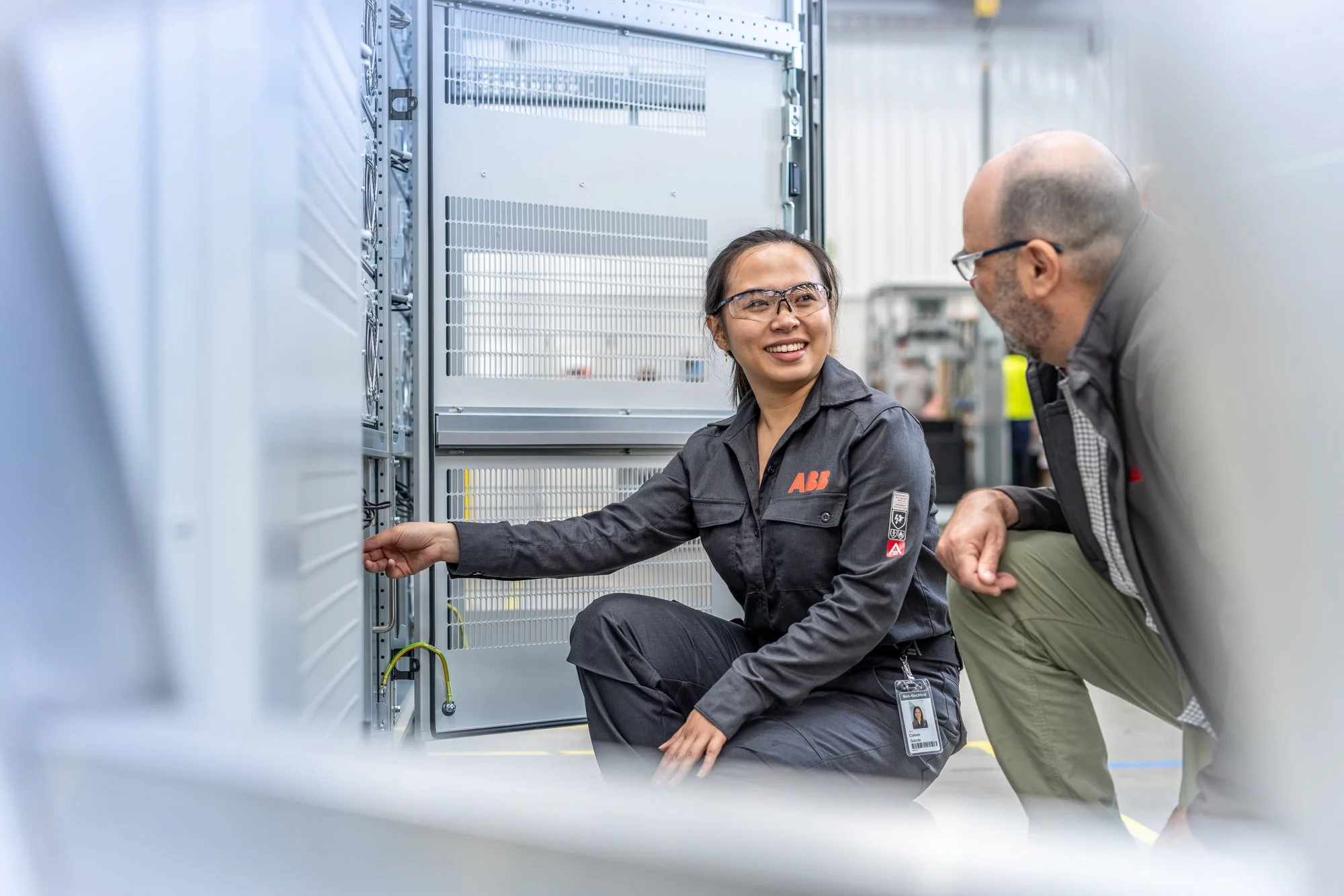 A woman in safety glasses and a dark work uniform with an ABB logo is showing a man in safety glasses and a dark jacket a piece of equipment inside an industrial control panel or electrical cabinet, in a manufacturing or service facility.
