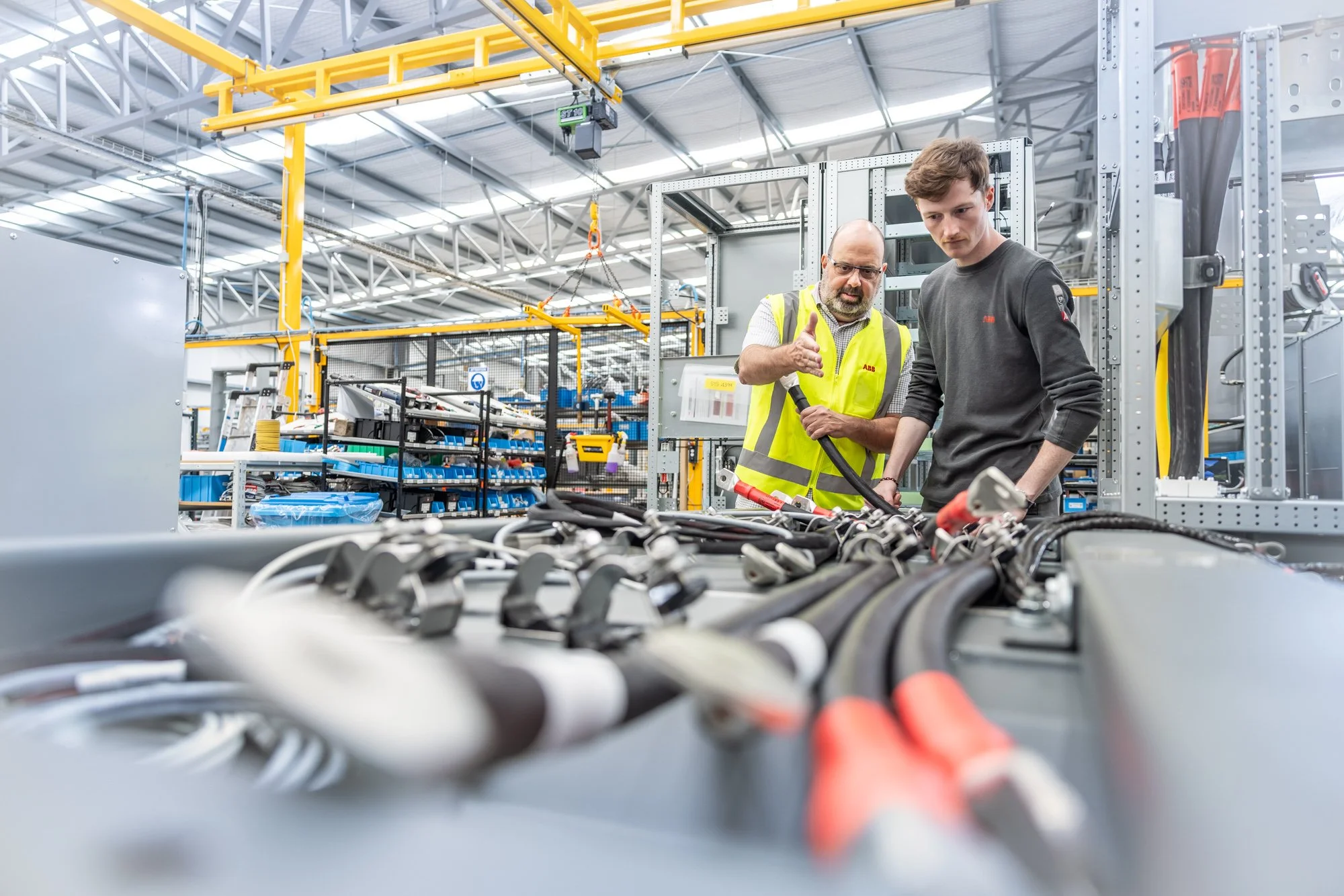 Two workers, one in a yellow safety vest and the other in dark clothing, inspecting and working on electrical cables or wires in an industrial warehouse or manufacturing facility.  Photographed by Alex Wallace Photography