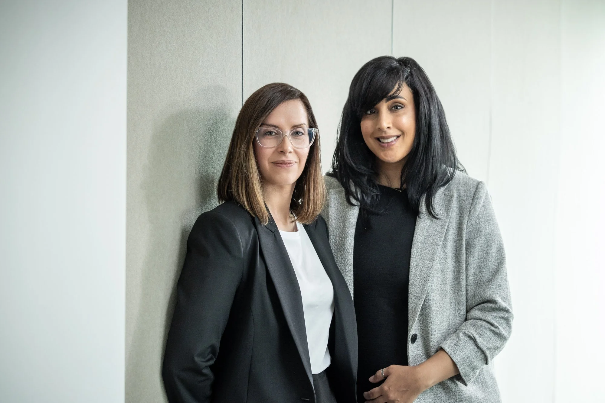 Two women standing side by side, smiling, against a plain wall. The woman on the left has shoulder-length brown hair, glasses, and is wearing a black blazer over a white top. The woman on the right has long black hair, and is wearing a grey blazer ov