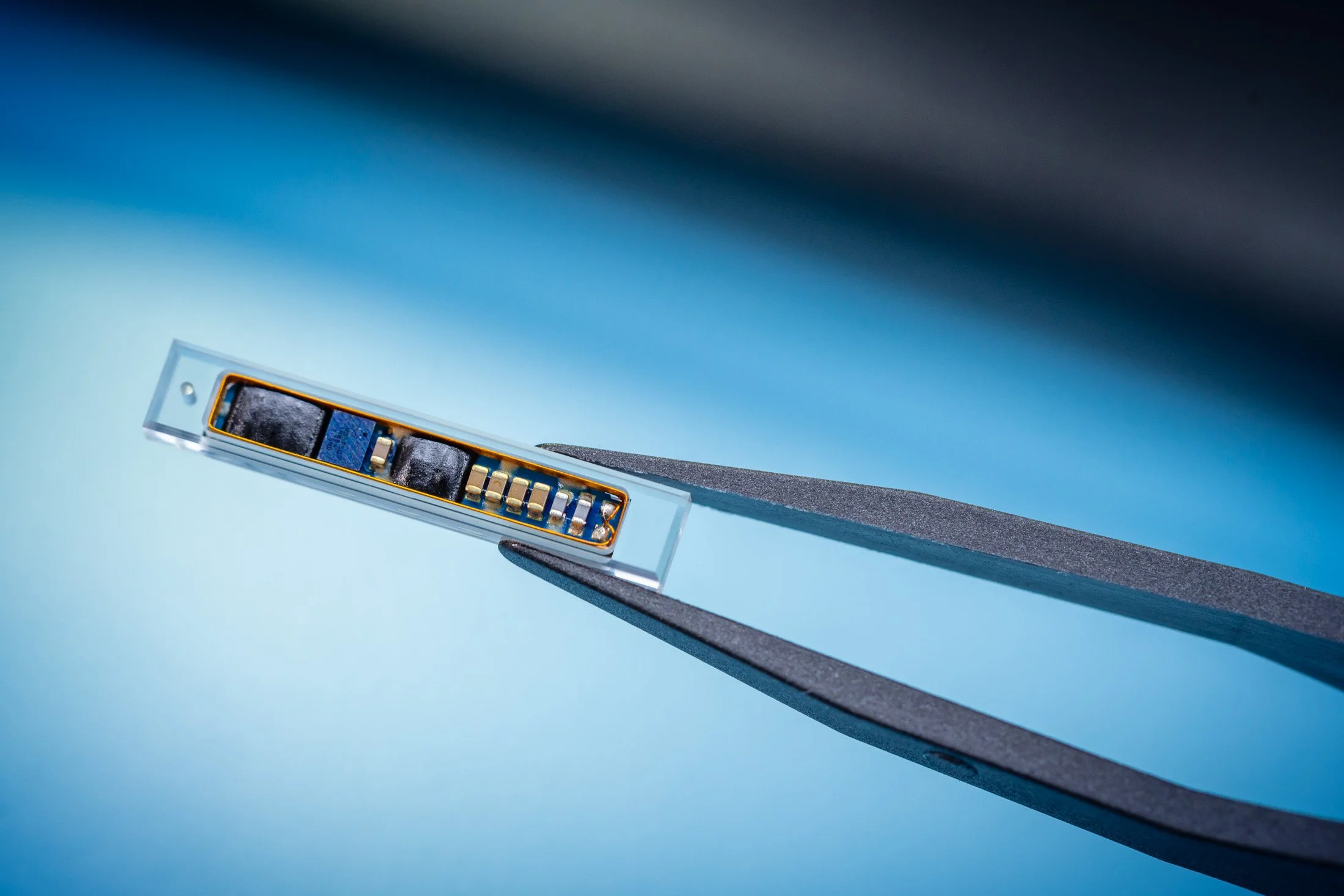 Close-up of a small electronic component held by tweezers on a blue gradient background.
