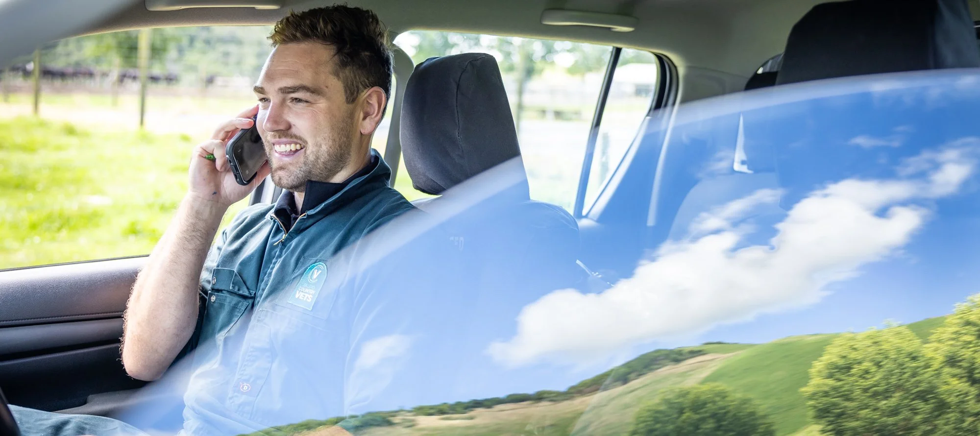A man smiling and talking on a cellphone inside a car, with a reflection of the sky and green landscape visible on the window.  Photographed by Alex Wallace Photography