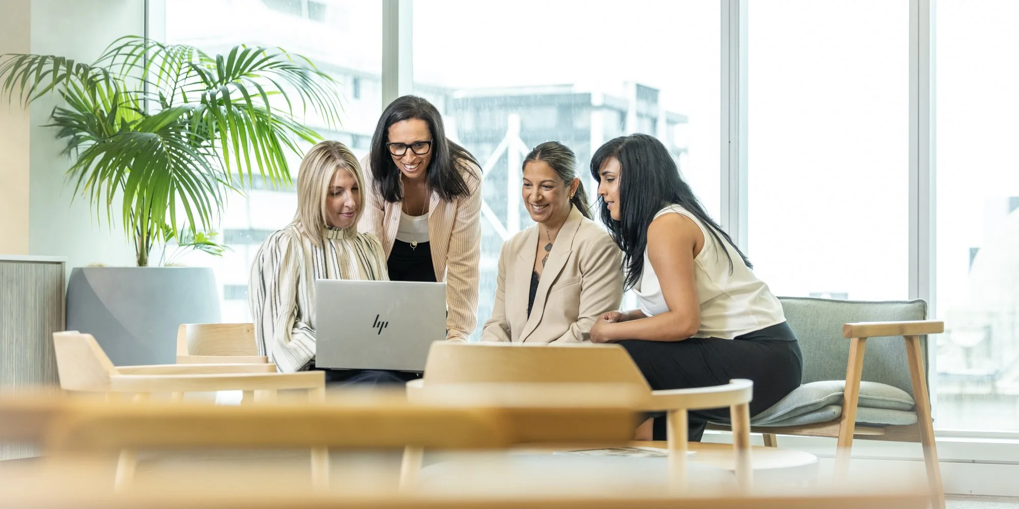 Four women gathered around a laptop in a bright office with large windows and a potted plant, smiling and engaged in a discussion.