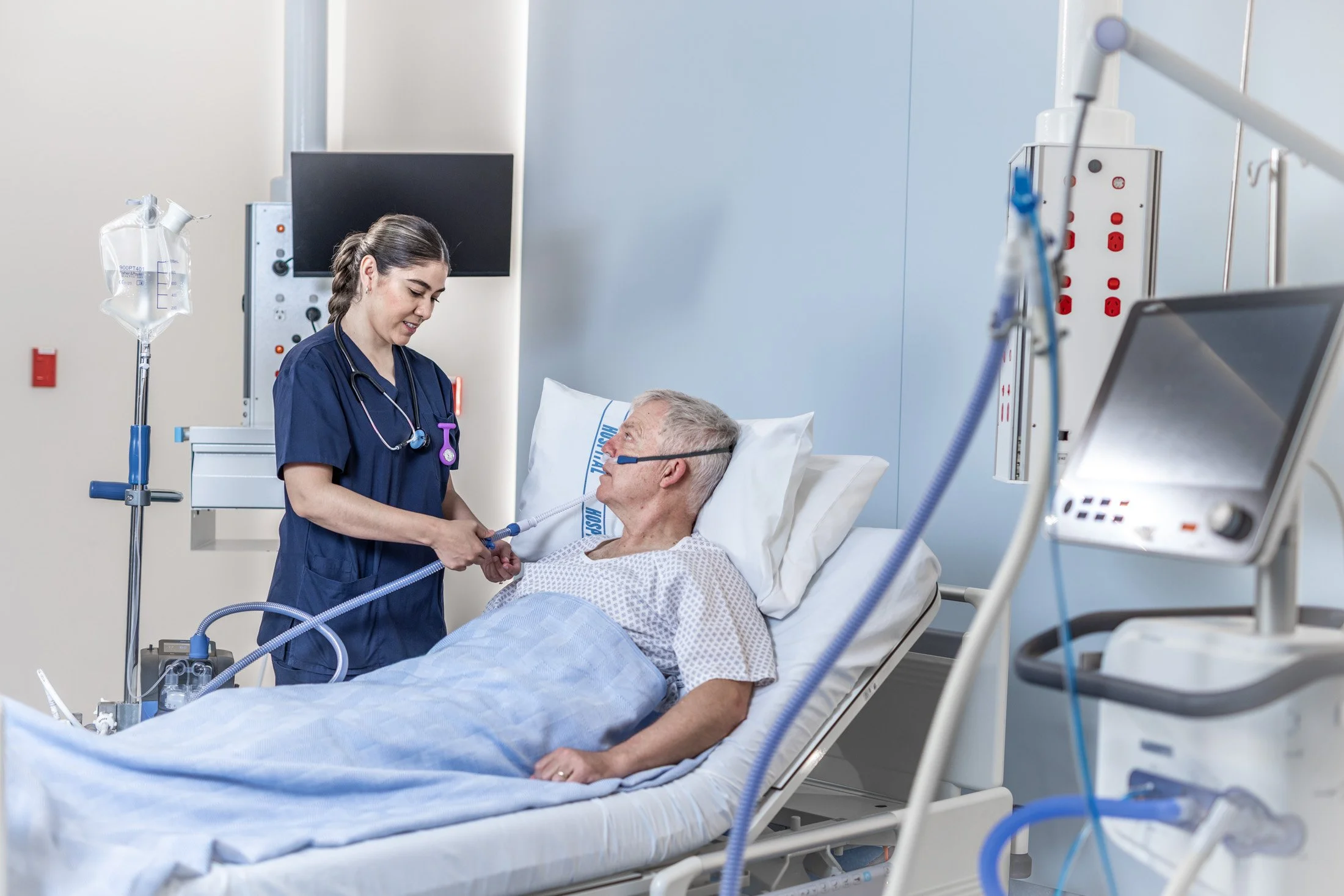 A young female nurse checks on an older male patient in a hospital bed, who is hooked up to medical equipment including a ventilator.