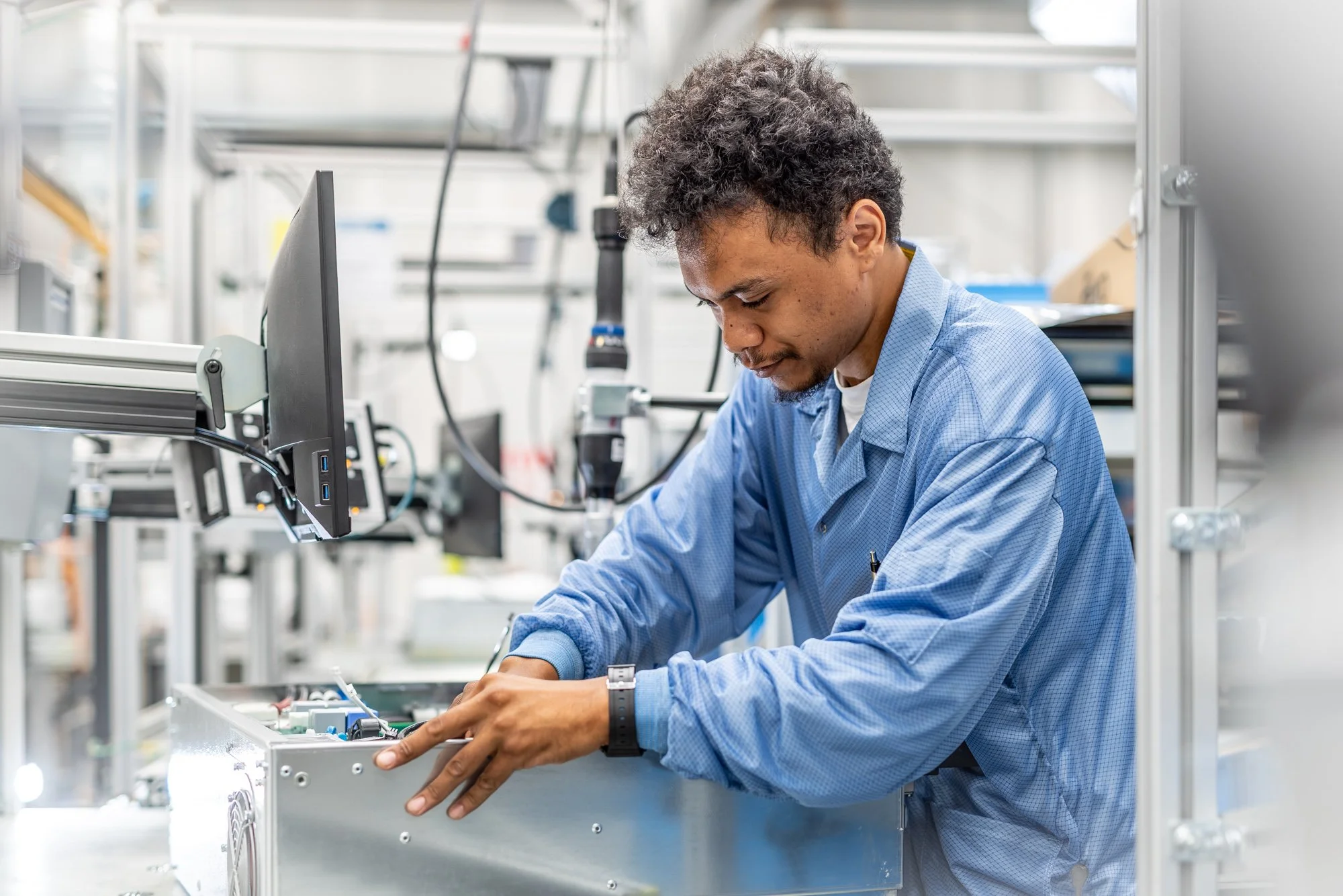 A male technician wearing a blue lab coat works on an electronic device inside a laboratory or manufacturing facility with shelves and equipment in the background.