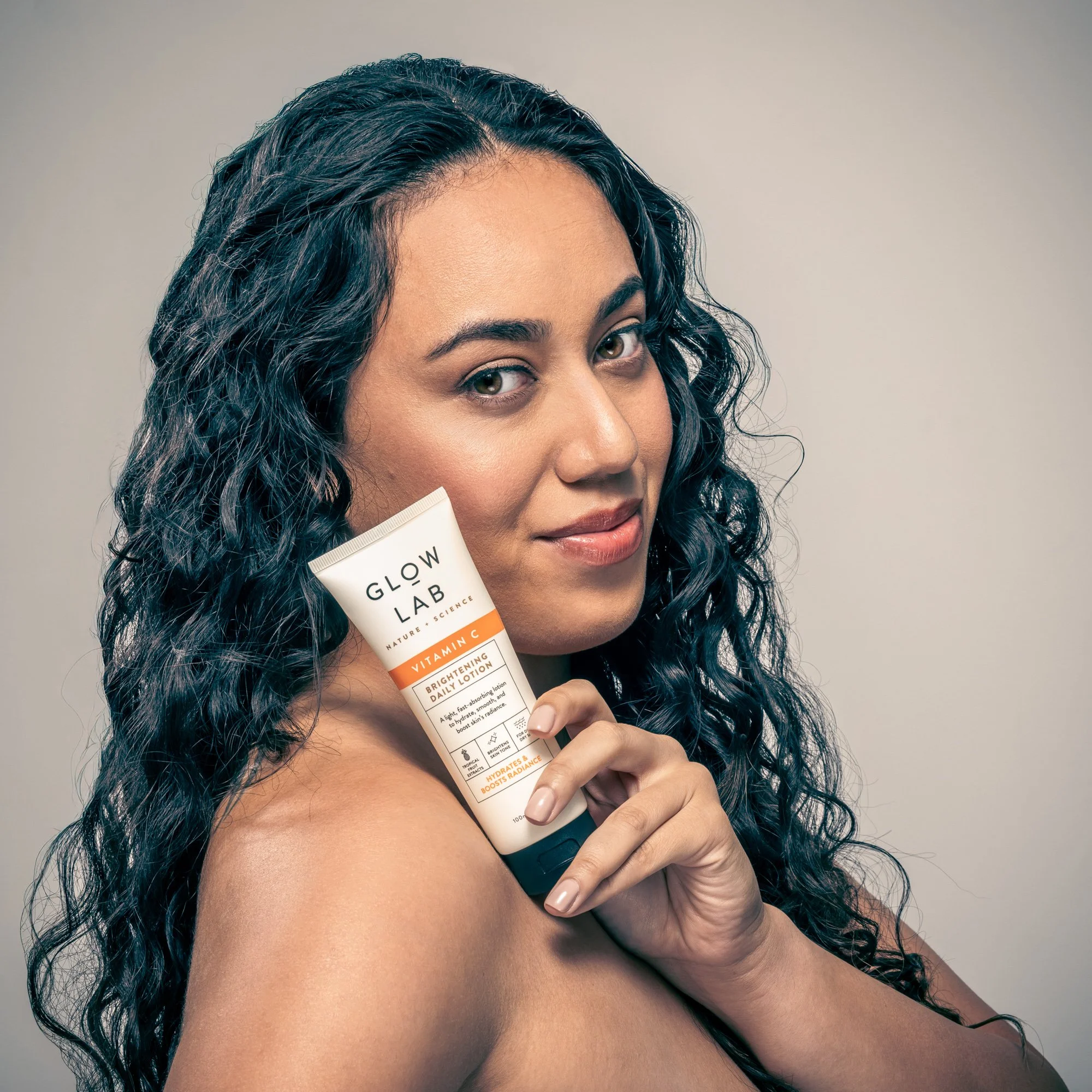 A woman with long dark curly hair holding a tube of Glow Lab vitamin C brightening daily lotion near her face, smiling softly against a neutral background.