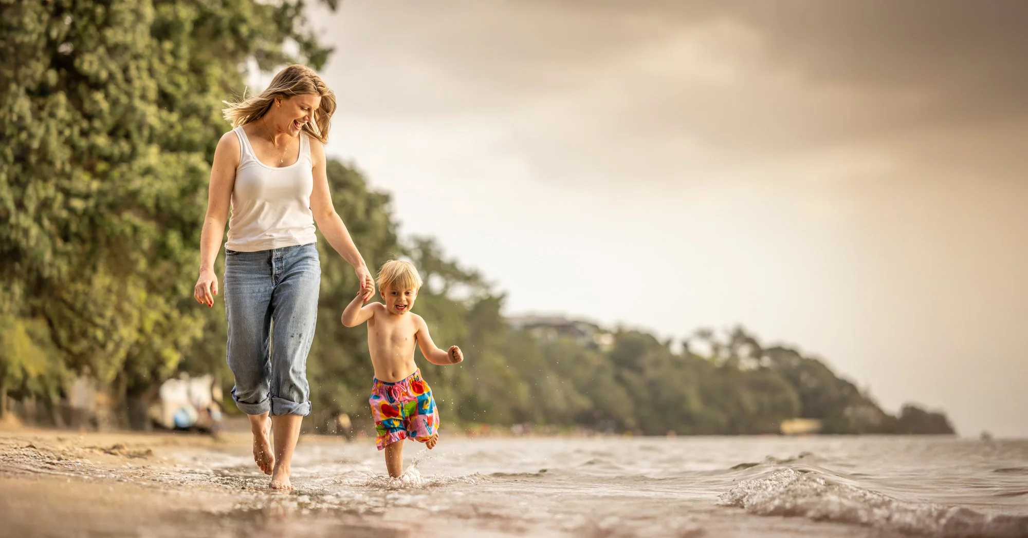 A woman and young boy walk barefoot along the shoreline of a beach, holding hands and smiling, with trees and a cloudy sky in the background.