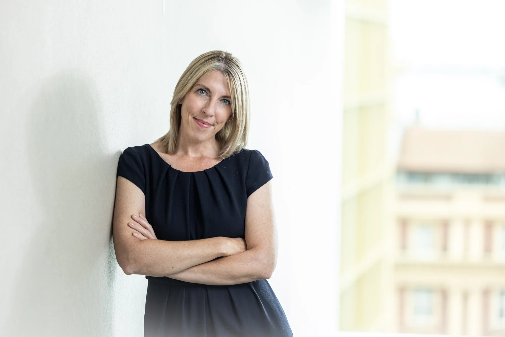 A woman with blonde hair wearing a black dress, standing with arms crossed and leaning against a wall in a well-lit indoor setting.
