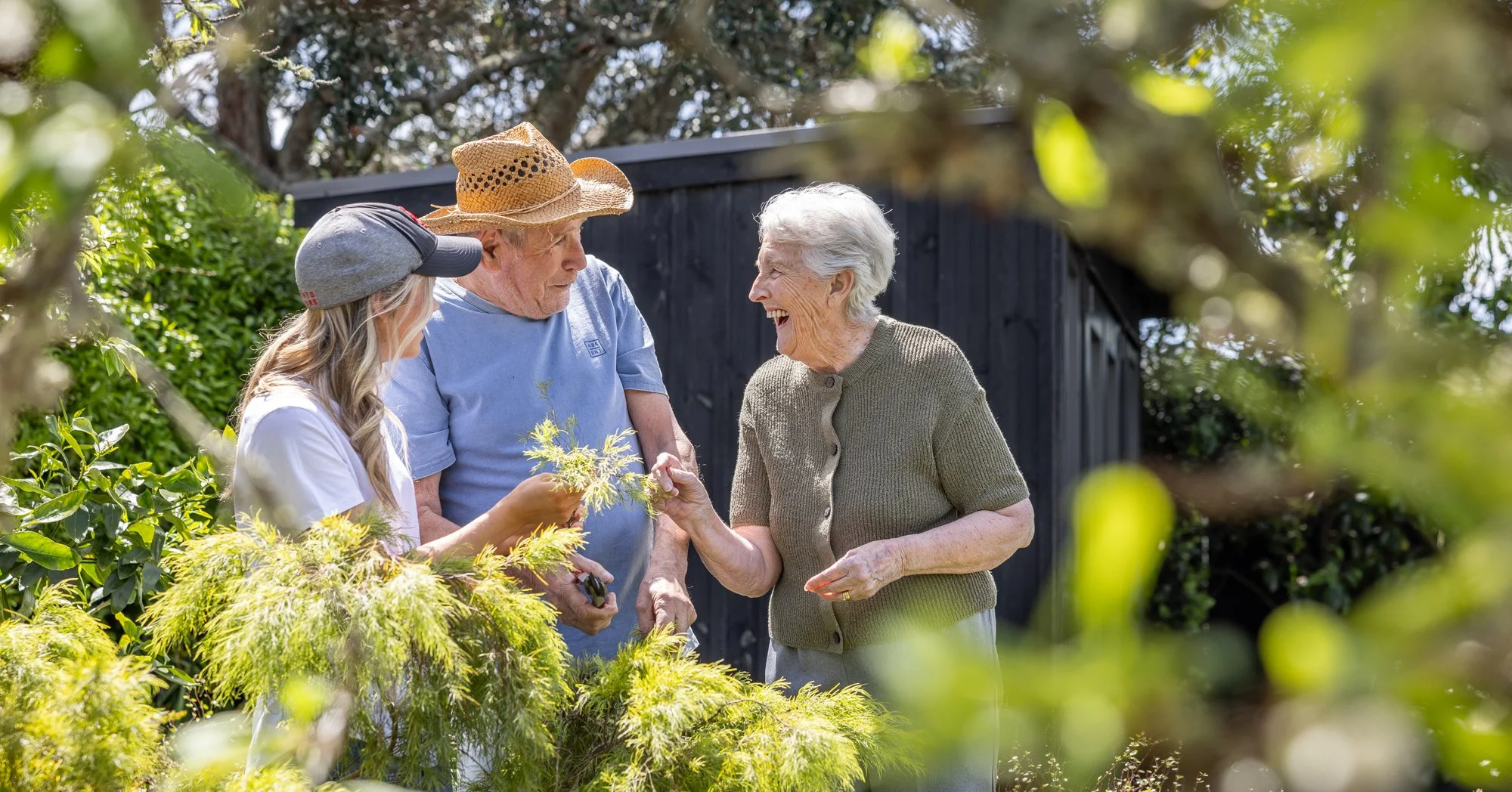 Three generations of women gardening together outdoors, with greenery and a black shed in the background. The elderly woman smiling, the middle-aged woman holding a plant, and the young woman in a cap are engaged in a joyful conversation.