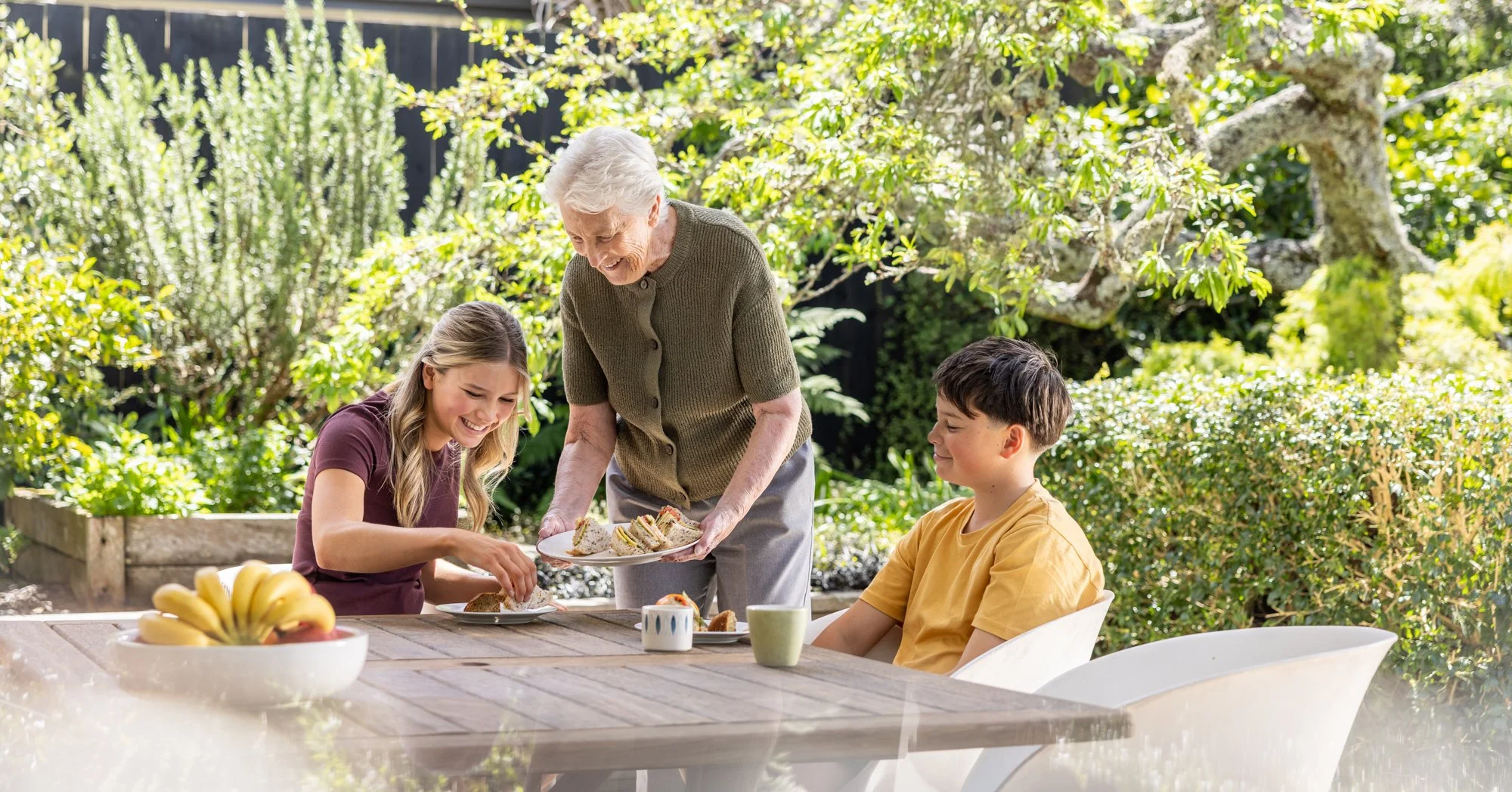 Grandmother serving tea and sandwiches to two children during outdoor breakfast in garden.