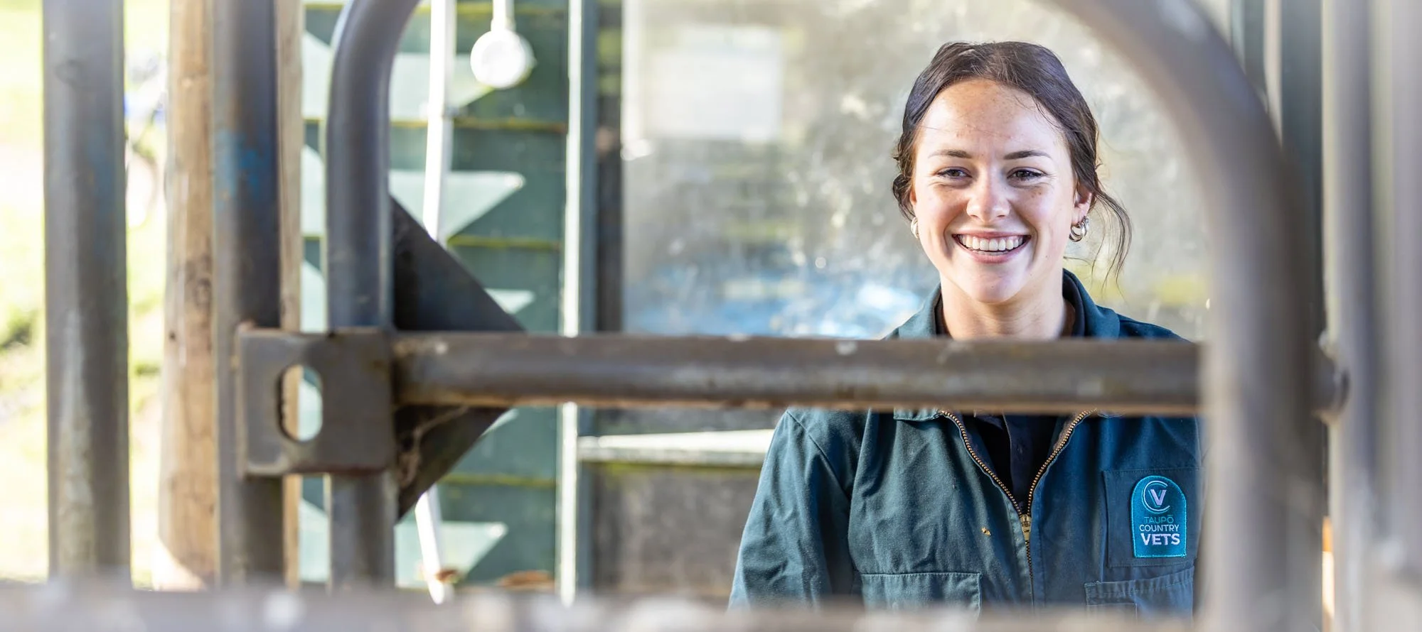 A woman with dark hair and earrings smiling through a metal gate. She is wearing a dark jacket with a patch that says 'Vets' and 'Taupo County'.  Photographed by Alex Wallace Photography