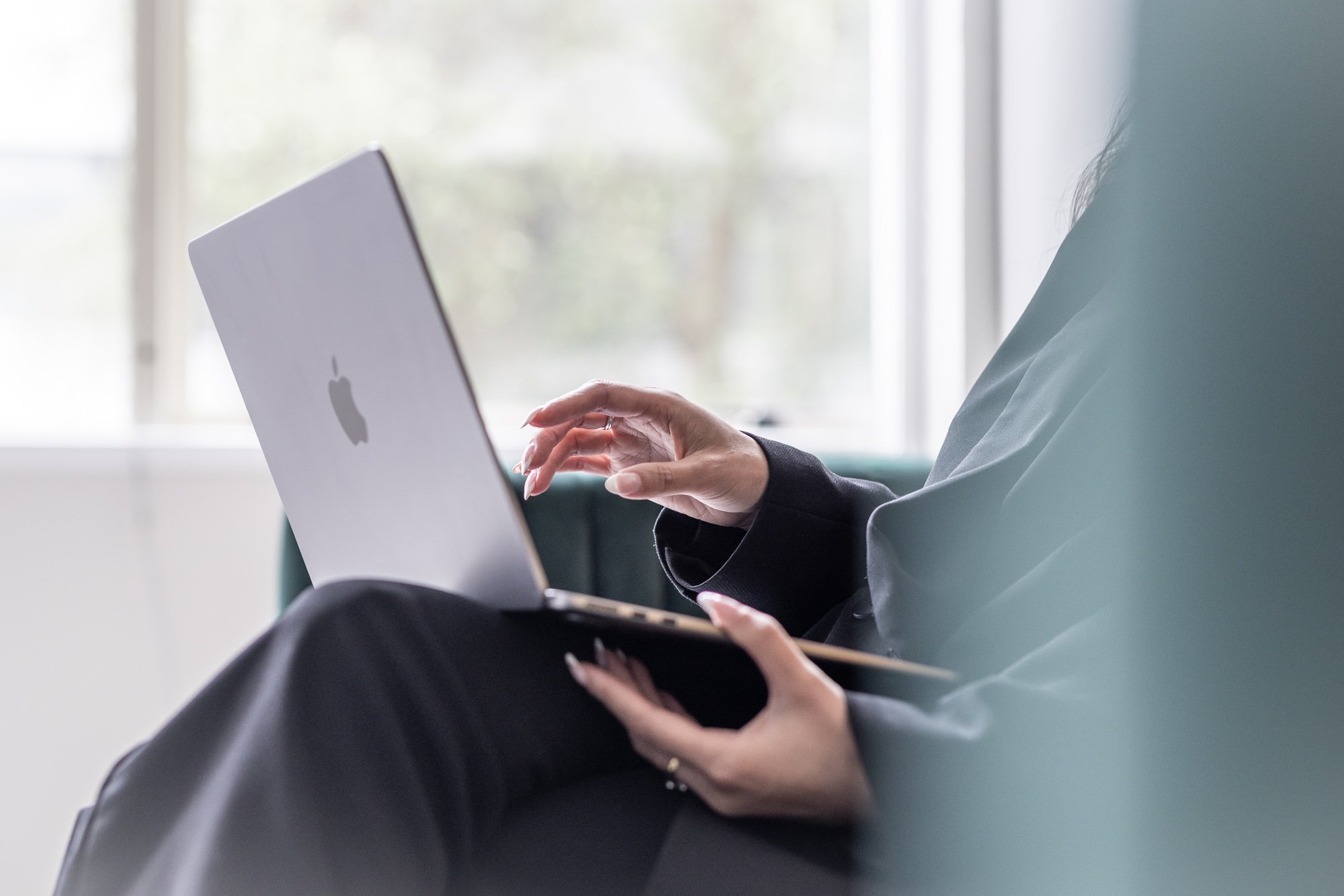 Person sitting on a couch using a silver MacBook with a blurred window background.