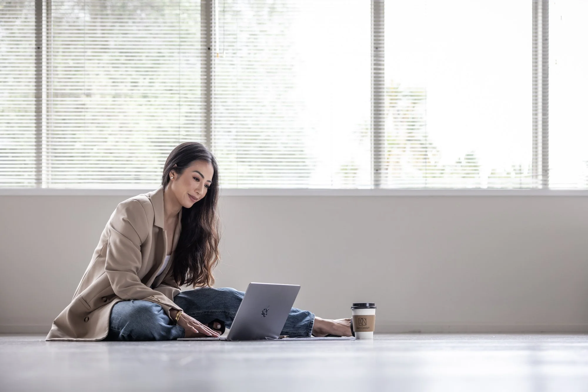 Woman sitting on the floor in a bright room, working on a laptop with a coffee cup beside her.