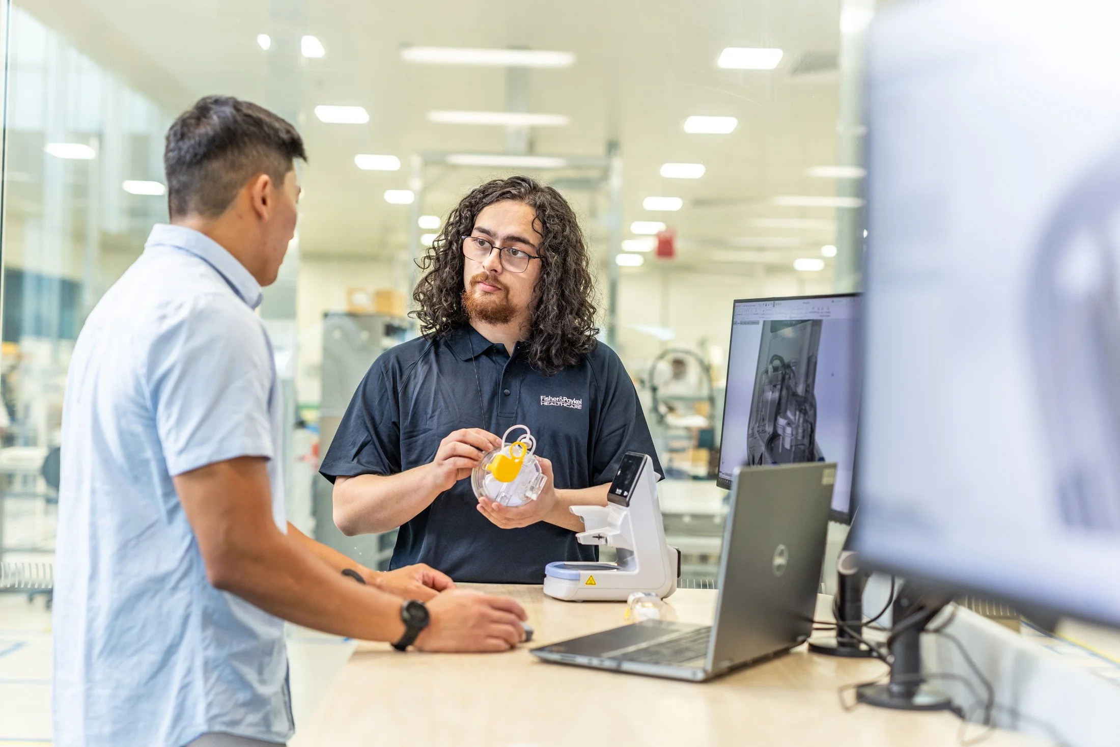 Two men in a laboratory, one explaining a medical device to the other. One is wearing a light blue shirt, and the other is wearing a dark polo with a logo. They are standing at a desk with a laptop, a 3D printer, and a computer monitor in a medical o