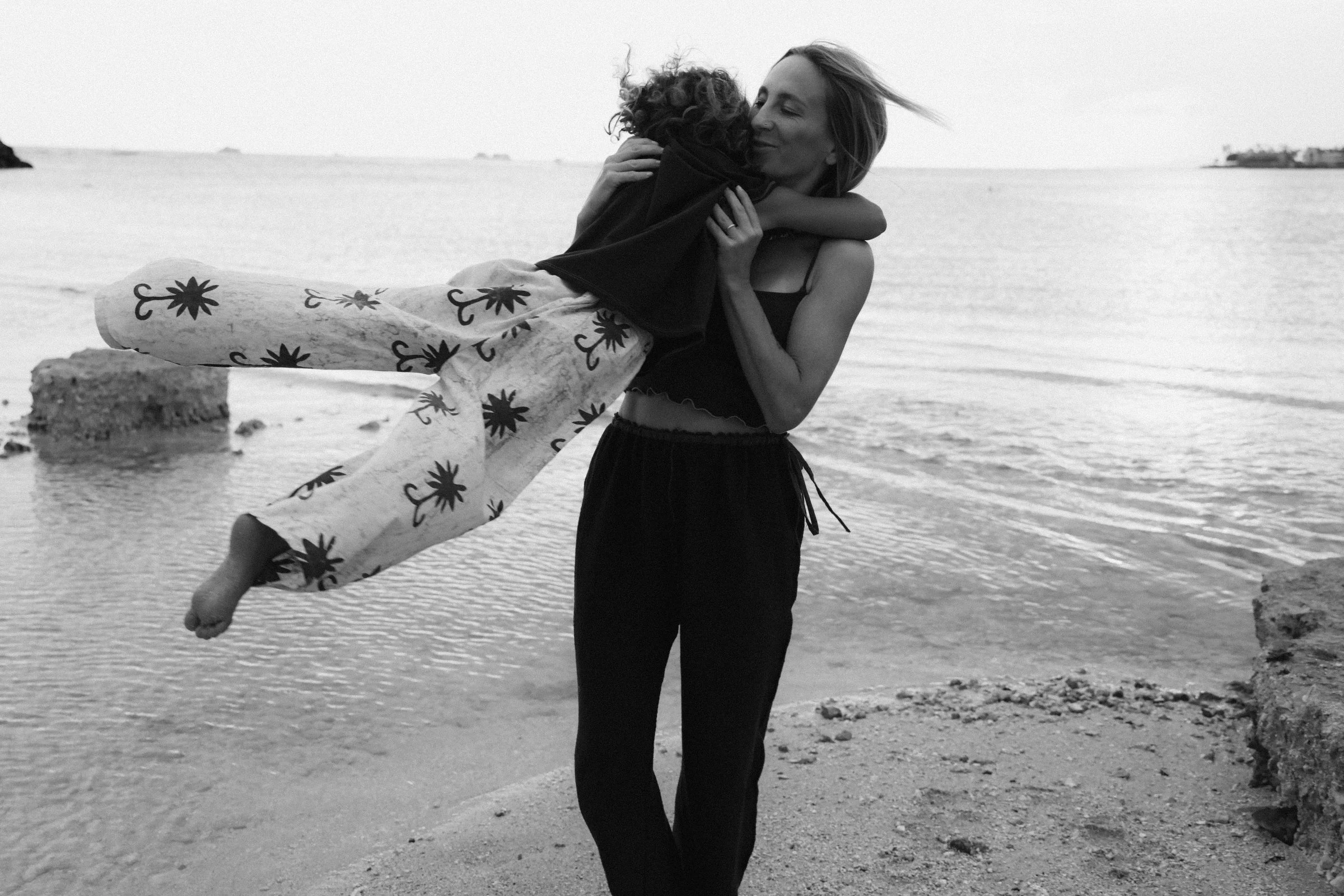 A woman holding a child on the beach near the water, embracing and smiling with the child. The scene is black and white.