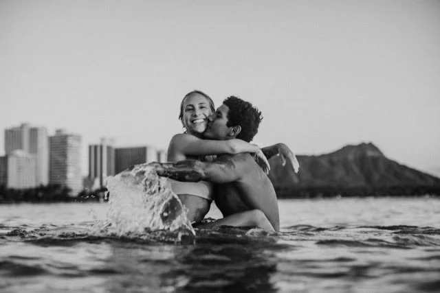 A couple hugging in the water at the beach with a city skyline and mountains in the background.