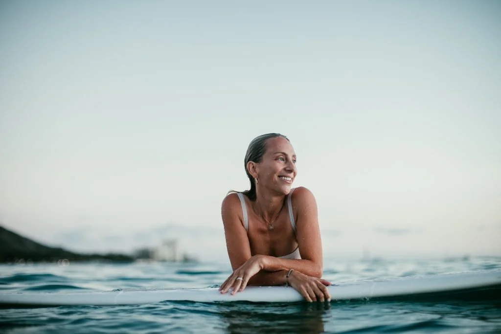 A woman smiling and sitting on a surfboard in the water during sunset or sunrise.