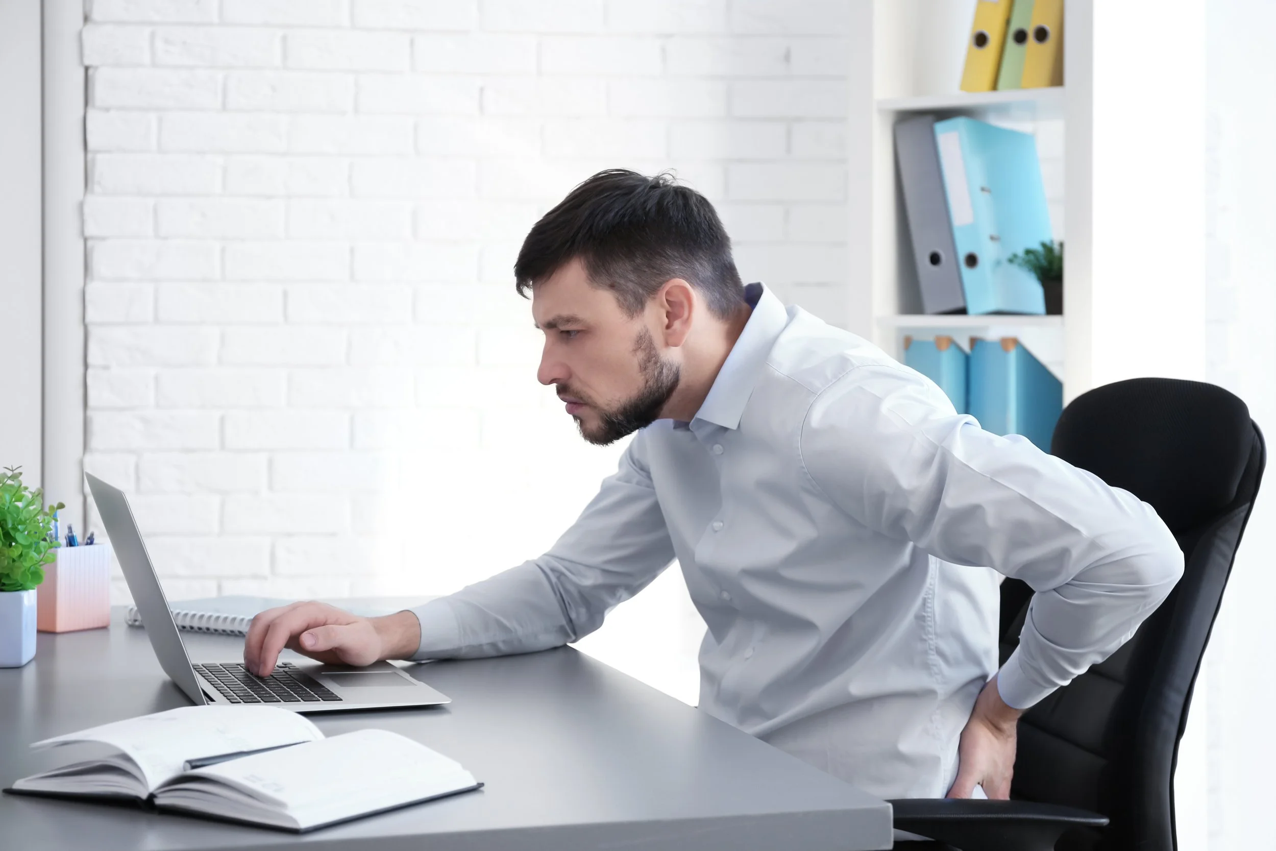 Man holding back pain, sitting at desk, looking at laptop screen
