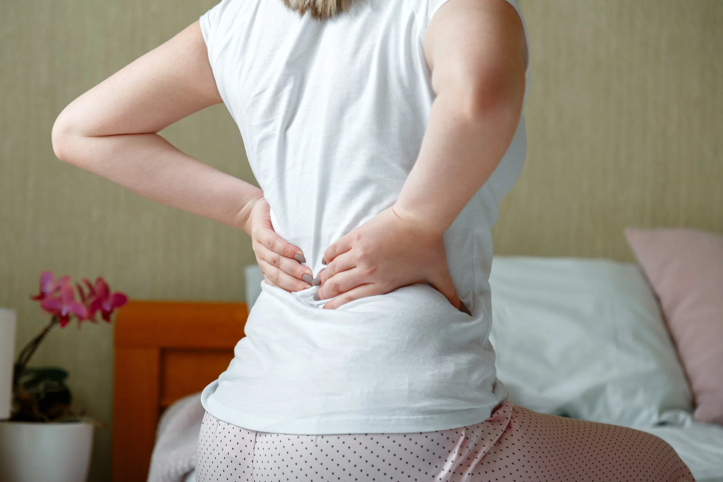 Person with back pain sitting on a bed, holding lower back, wearing a white shirt and polka dot pants, with blurred flowers in the background.