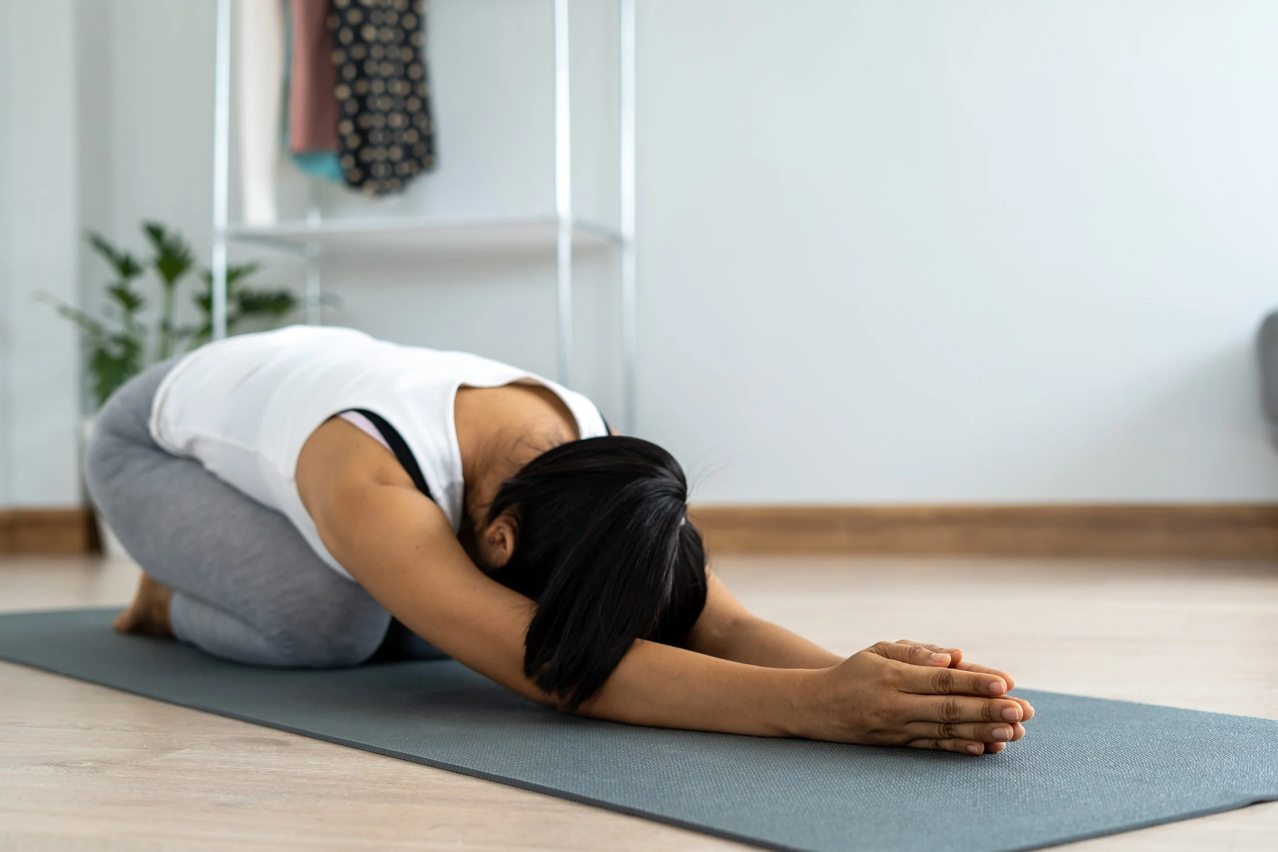 A young woman in exercise wear sitting on yoga mat on the floor leaning forward with her arms stretched