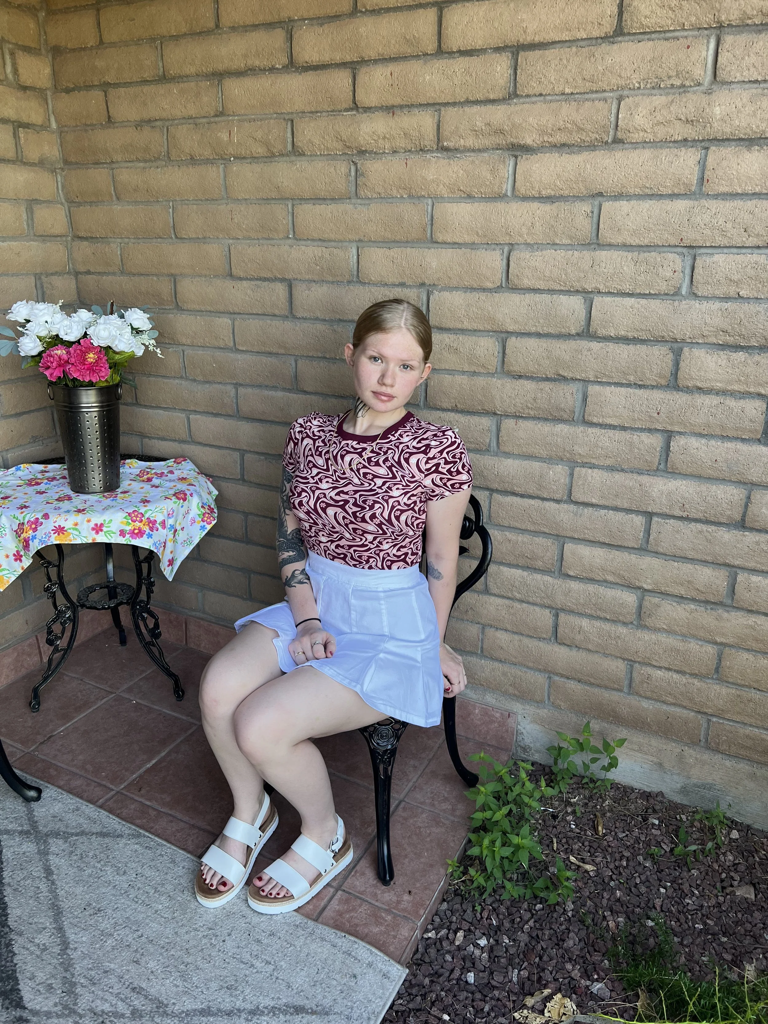 A person sitting on a chair by a brick wall. They are wearing a patterned top, a light-colored skirt, and white sandals. Beside them is a small table with a floral tablecloth and a vase of flowers. Co owner of Legends AZ.