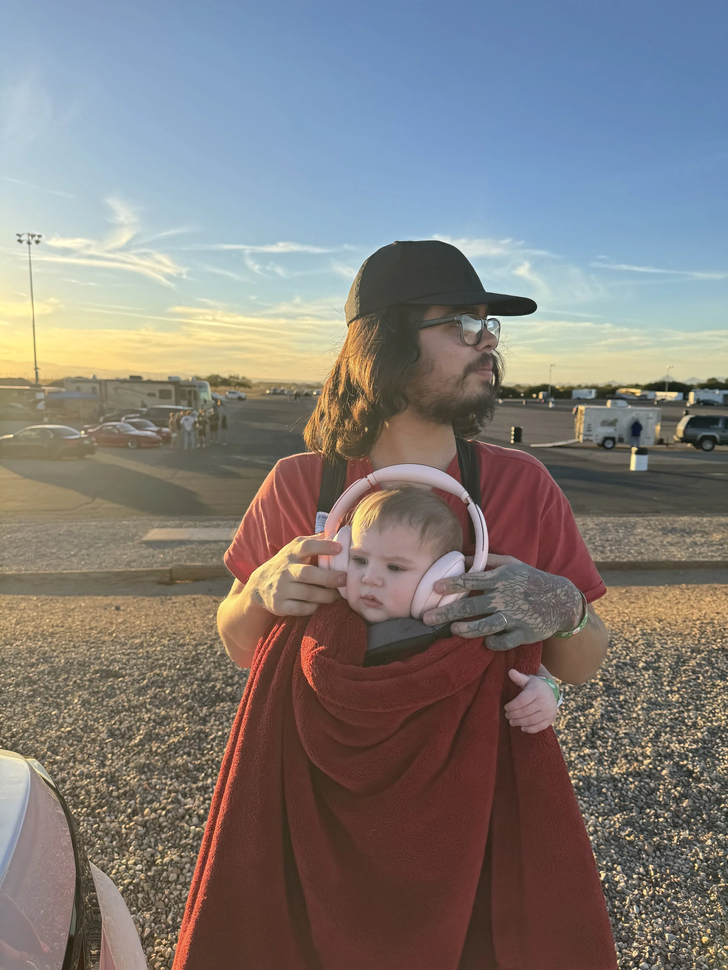 Person holding a baby wrapped in a red blanket with headphones, outdoors with a sunset in the background. Owner of Legends AZ.