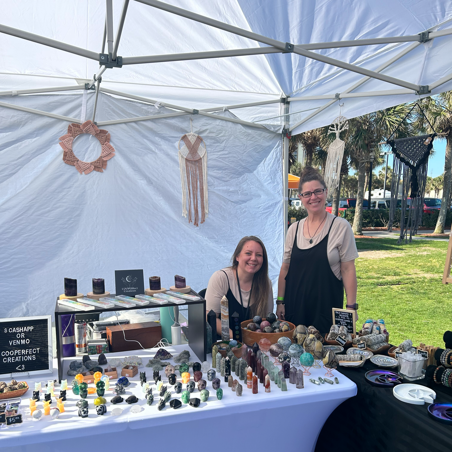 Two women working at a crystal and jewelry booth at an outdoor market, with handmade decorations hanging above and palm trees in the background.