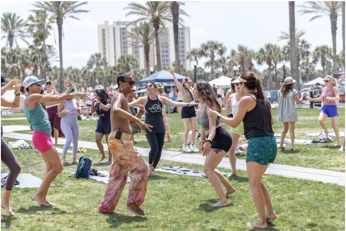 People dancing and having fun outdoors on a sunny day at a park with palm trees and a cityscape in the background.