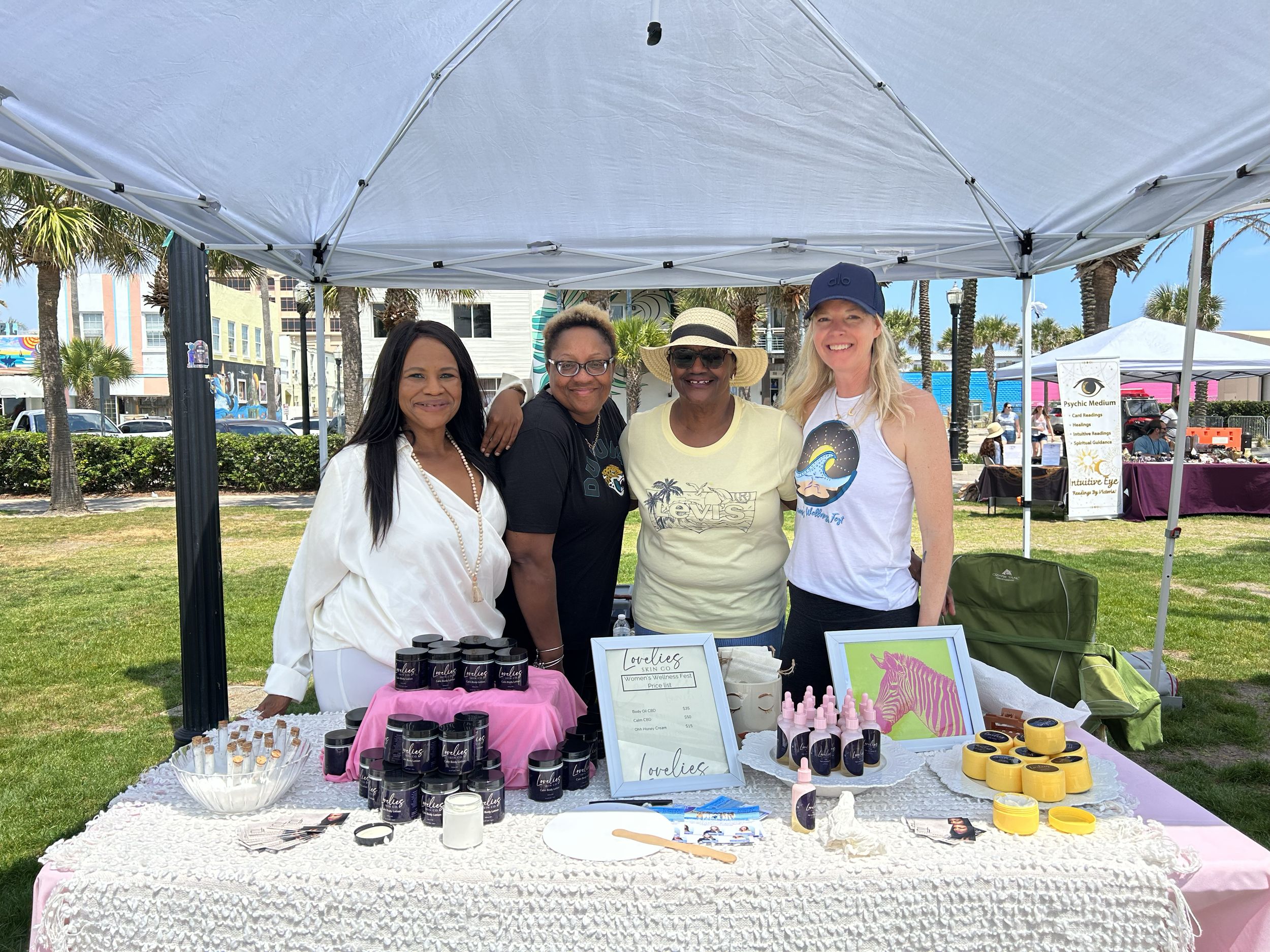 Four women standing behind a table with skincare and beauty products at an outdoor event under a white canopy, with palm trees and tents in the background.