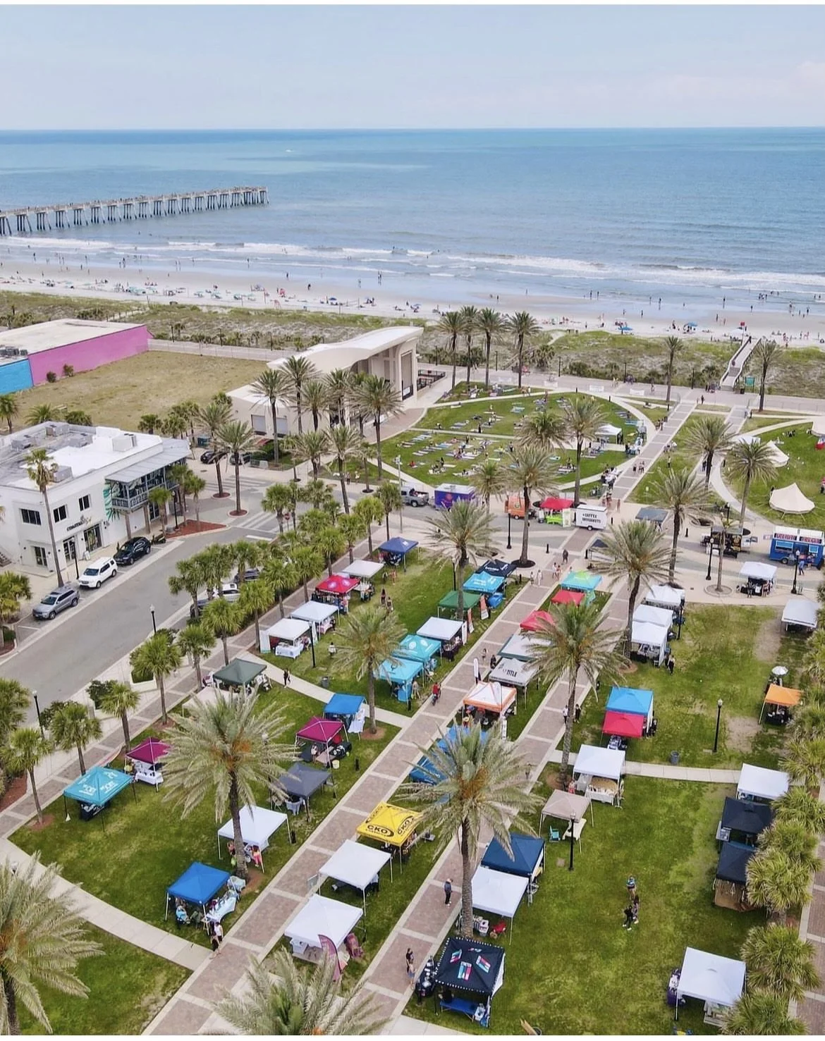 Aerial view of a beachside marketplace with colorful vendor tents, palm trees, a grassy area, and the ocean in the background.