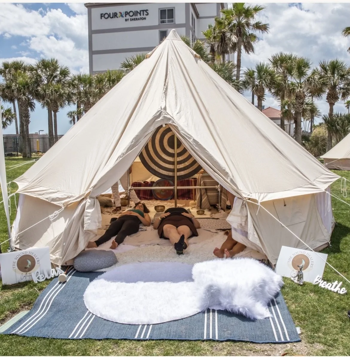Outdoor wellness event with a tipi tent set up on grass, featuring people relaxing inside with Indian-style bowls, surrounded by signs that say 'relax' and 'breathe', and decorative items like cushions and a fuzzy rug, with palm trees and a hotel building in the background.