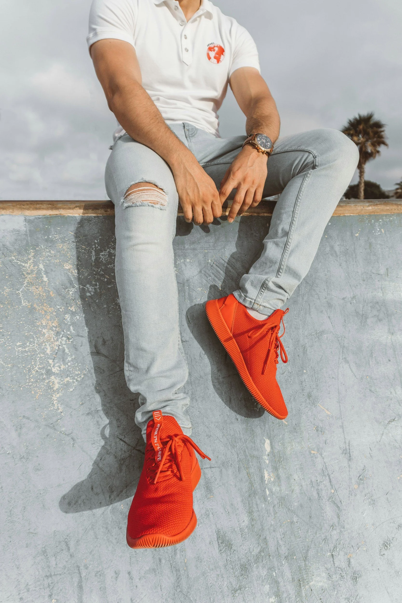 Man wearing bright red sneakers, light blue ripped jeans, and a white polo shirt sitting on a concrete ledge, representing the Men’s Streetwear section on the Aglo Designs merchandise page.