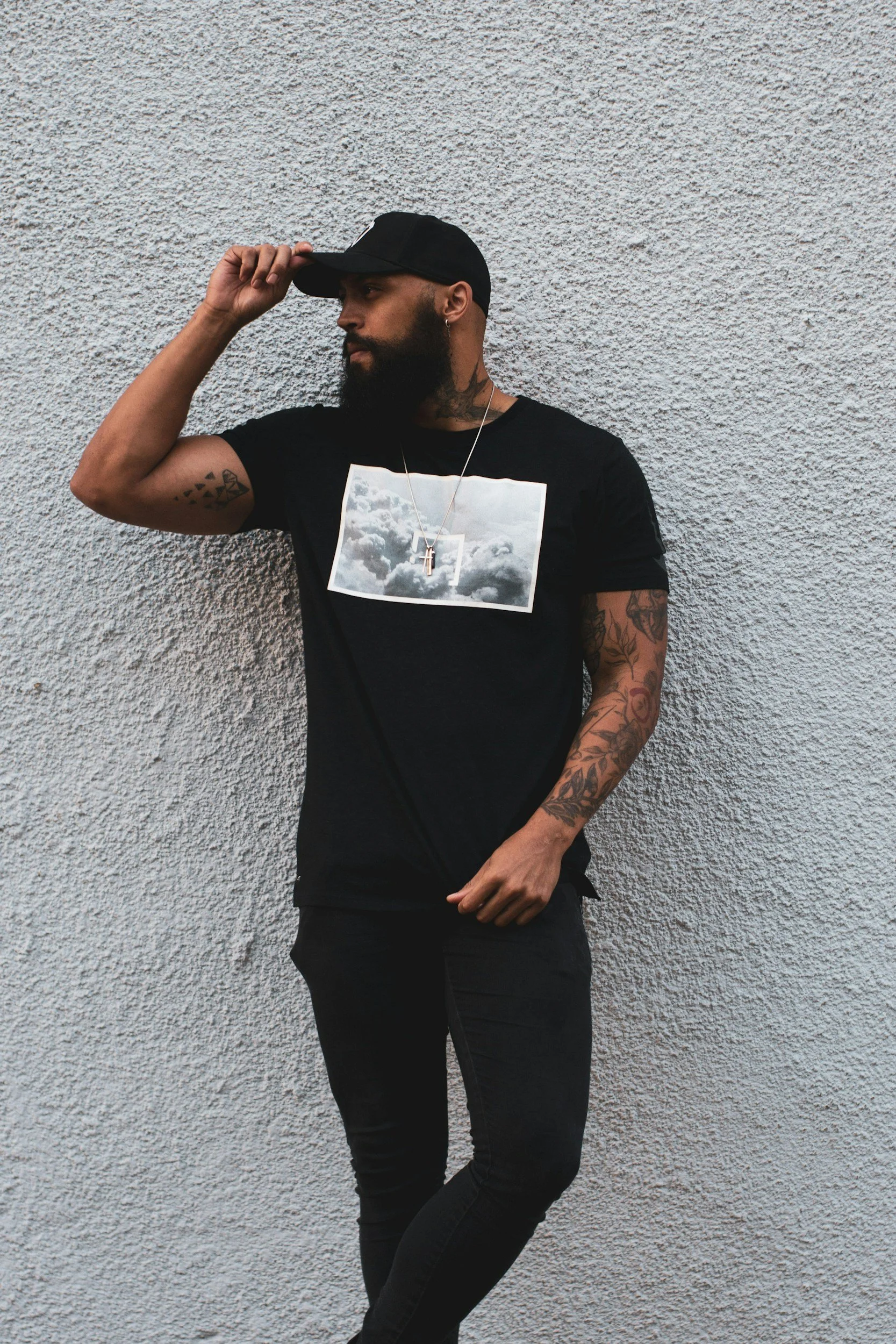 Man wearing a black cap and printed black T‑shirt standing against a textured gray wall, representing the Men’s Urban Collection on the Aglo Designs merchandise page.