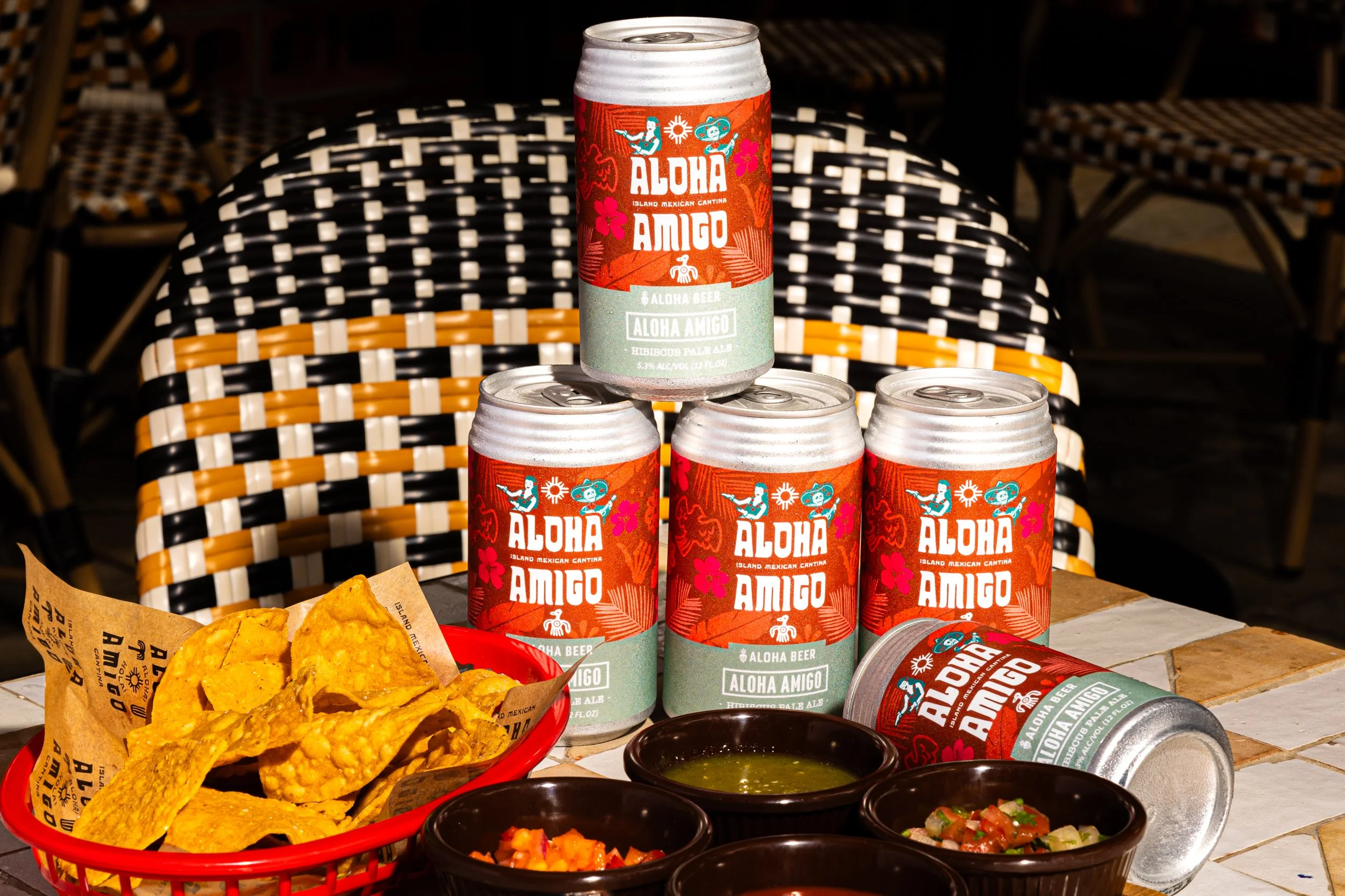 Four cans of Aloha Amigo beer with tropical design on a table, accompanied by bowls of salsa, chips, and green sauce, with woven chairs in the background.