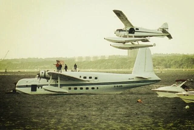 A beaver seaplane flying above a shorts Sunderland flying boat from WW2 on water, with a person carrying a flag and a few other people nearby.
