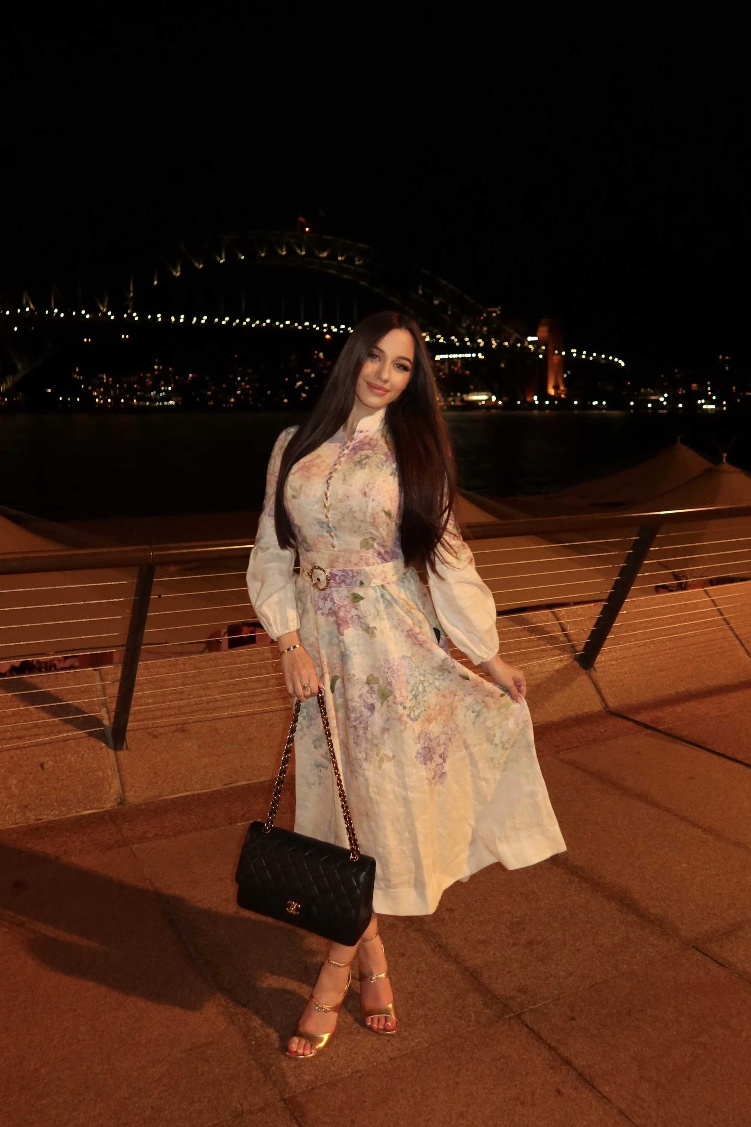 A woman in a floral dress and gold heels standing outdoors at night with a cityscape and bridge lit up in the background.