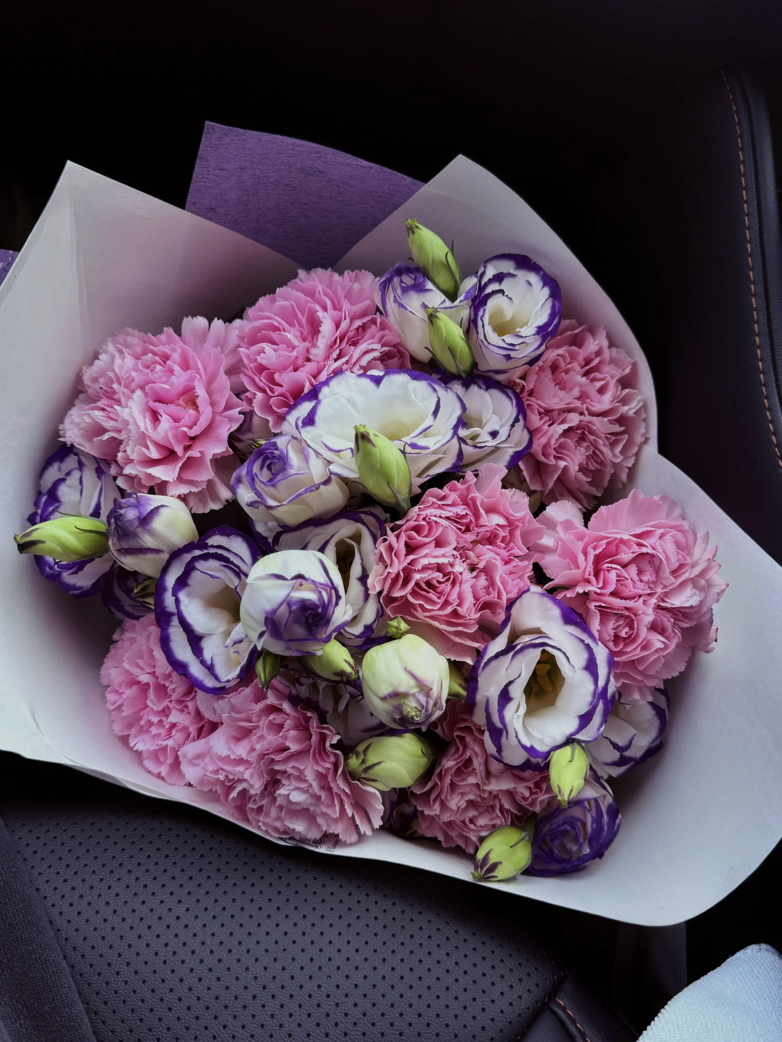 Bouquet of pink, purple, and white flowers on black car seat with orange stitching.