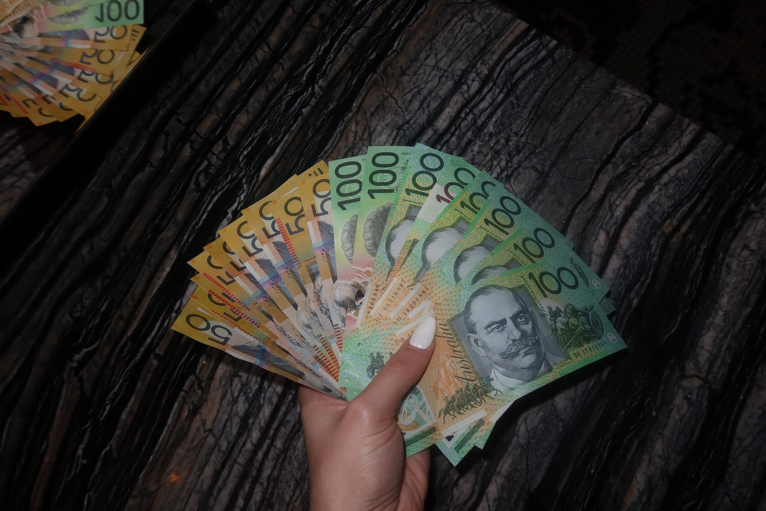Goddess Grace, Australian financial domination hand holding Australian currency from. a cashmeet in Sydney, Australia, including fifty dollar and one hundred dollar Australian notes, against a dark, textured surface.