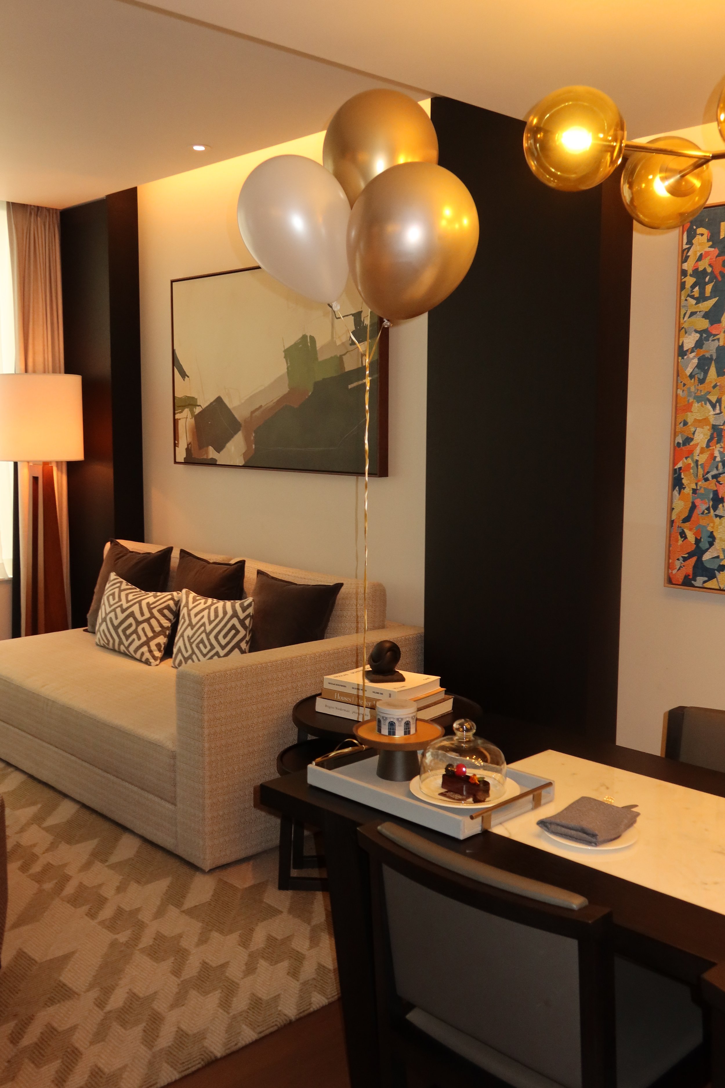 Findom funded suite in luxury hotel room with beige sofa, patterned pillows, black side tables, balloon arrangement with gold and white balloons, abstract wall art, and patterned beige rug.