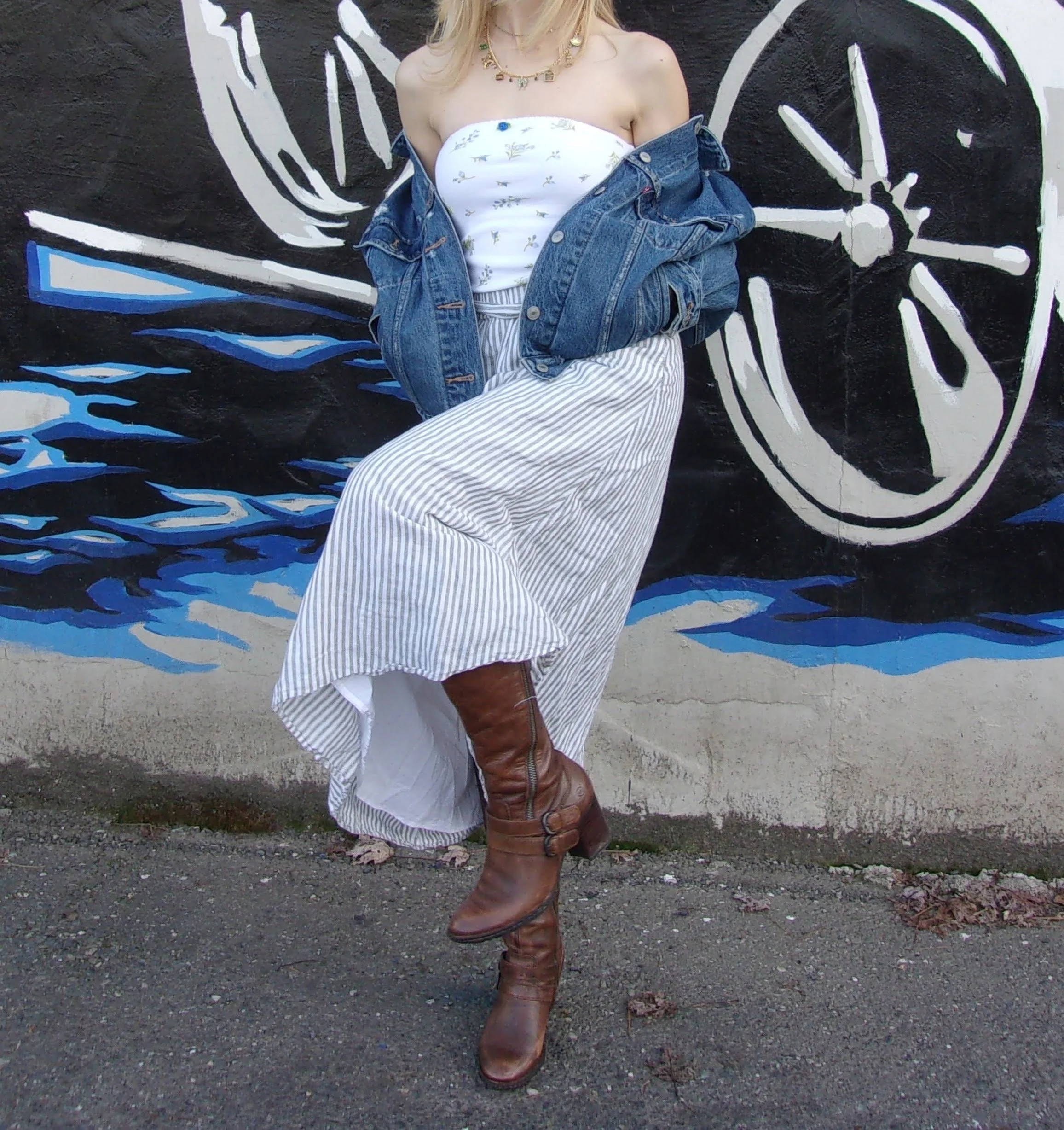 Person in a strapless white floral top, striped skirt, denim jacket off the shoulders, and brown knee-high boots standing in front of a black wall with a white and blue ocean wave mural.