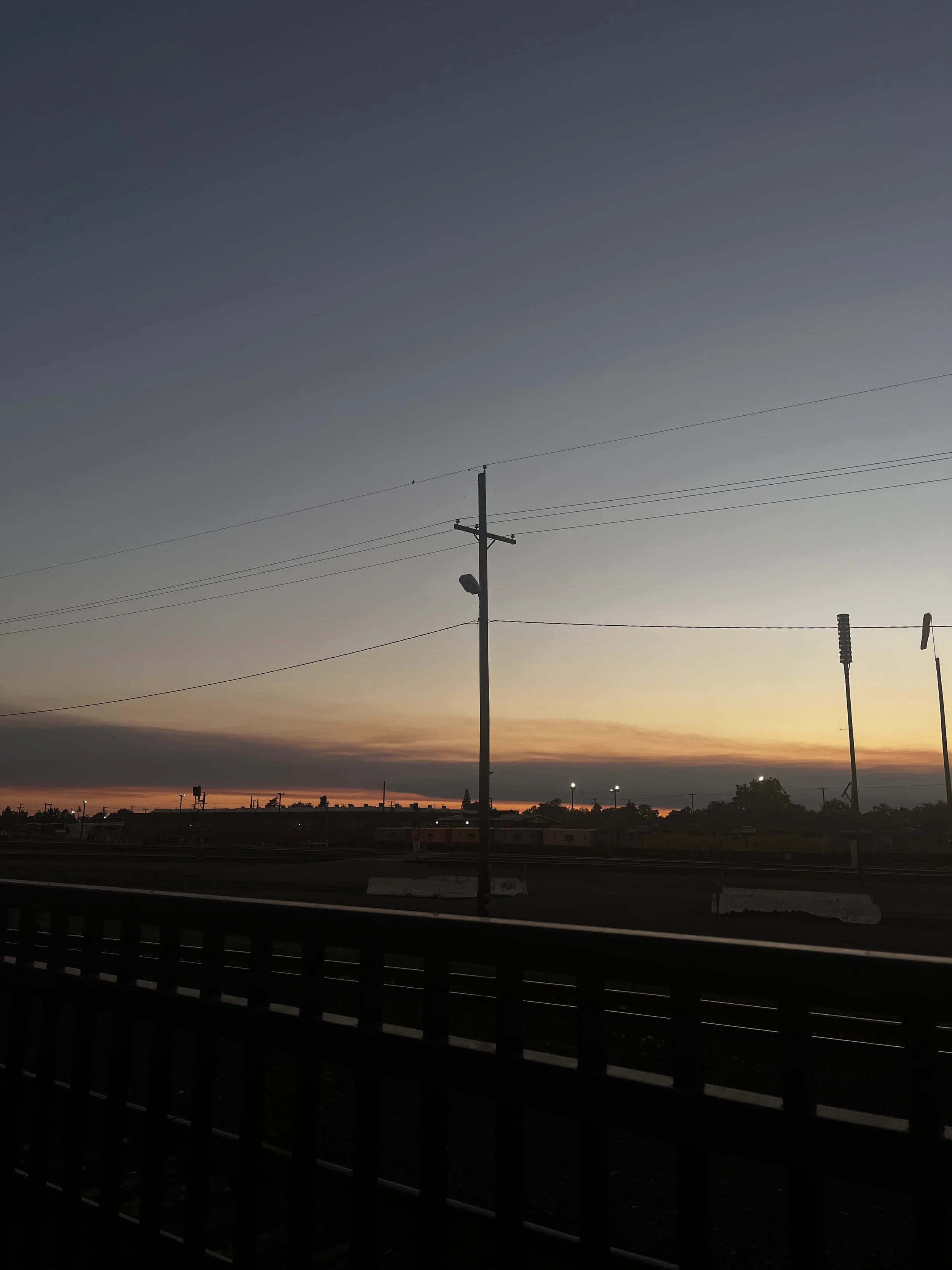 Sunset view over a train track with utility poles and power lines, with a fence in the foreground and distant city lights.