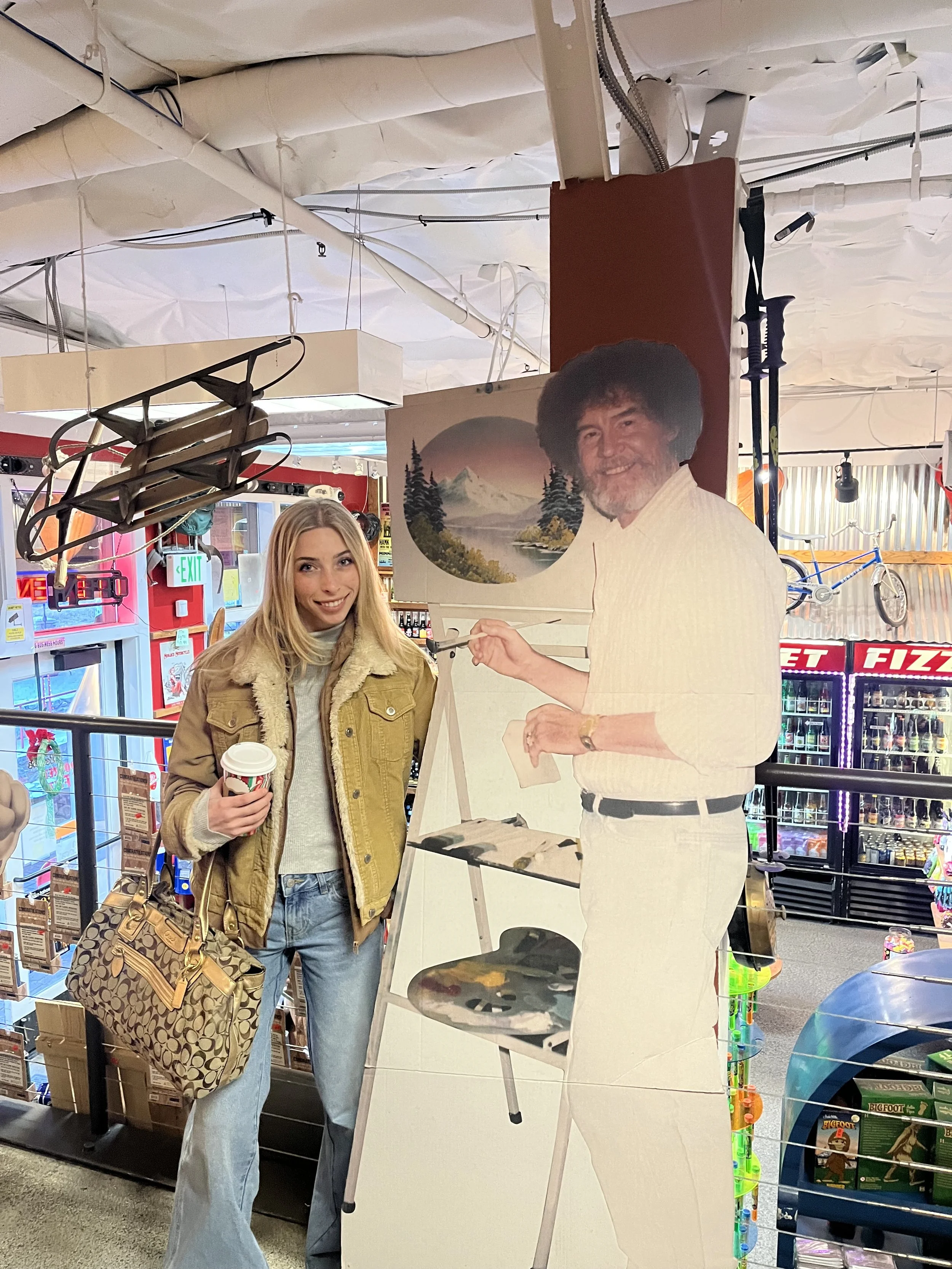 A young woman with blonde hair, wearing a brown jacket and light blue jeans, holding a coffee cup and a patterned handbag, standing next to a life-sized cardboard cutout of a man painting a landscape. They are inside a store with colorful shelves and beverages in the background.