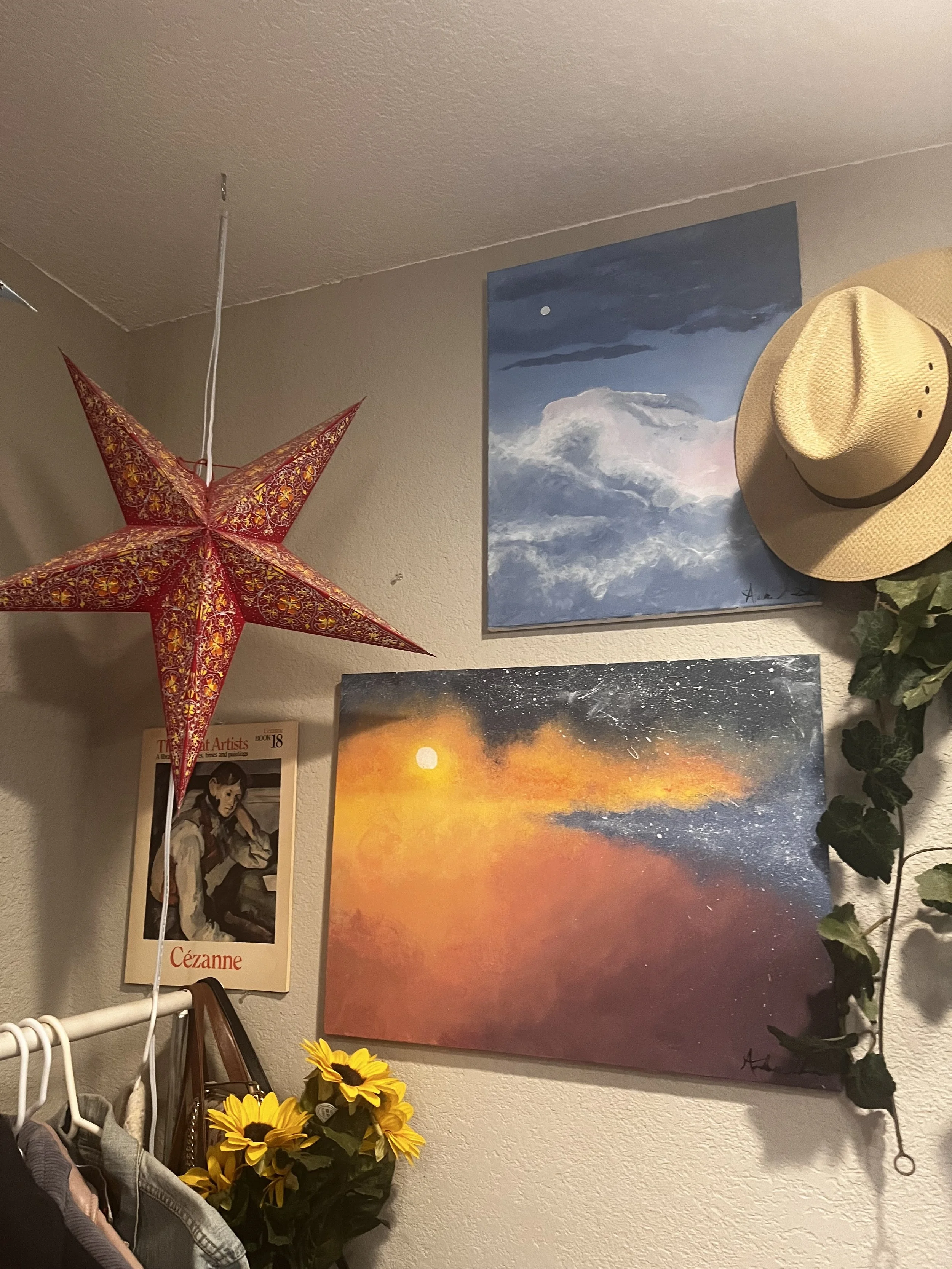 A hanging red and gold star-shaped lantern, two landscape paintings of clouds and sky, a straw hat, a poster of a woman of Asian descent, bunch of yellow sunflowers, and a green vine on a beige wall.